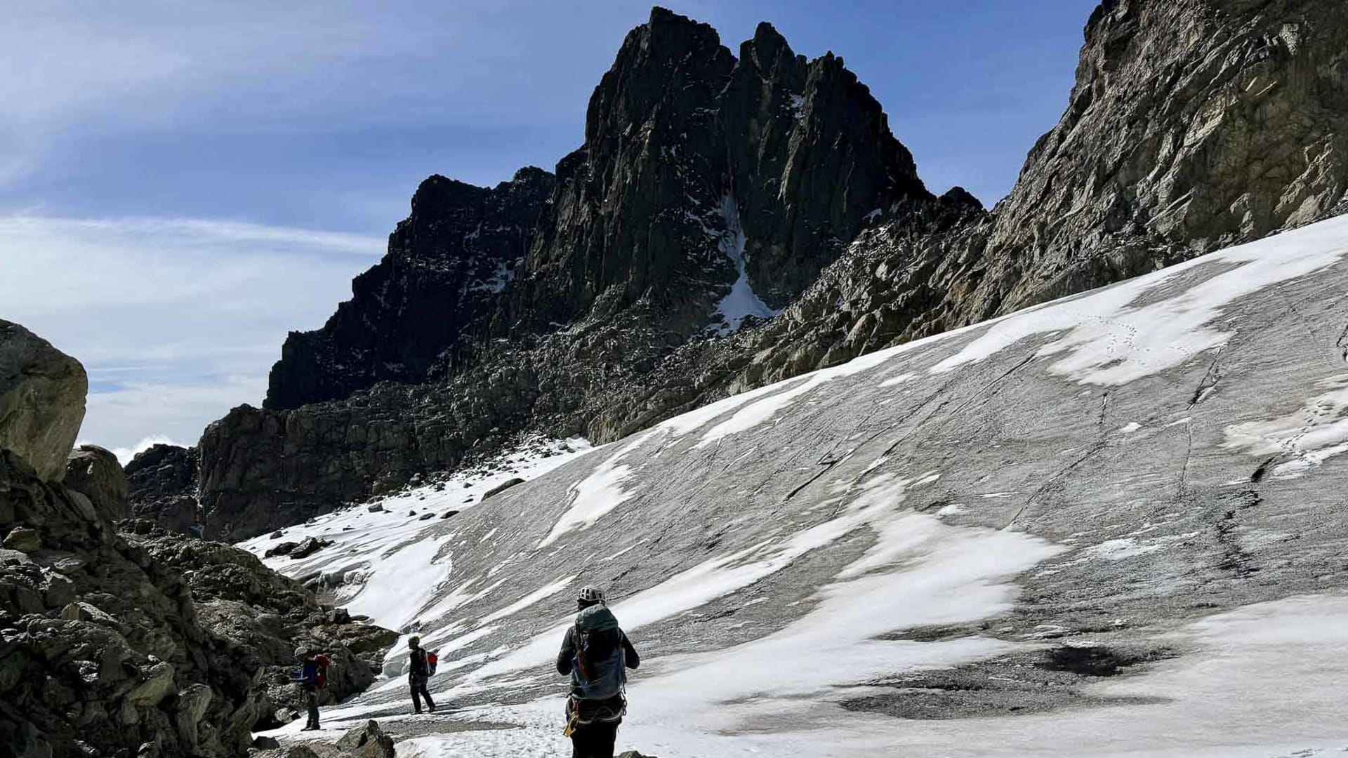 Drei Trekker mit Rucksäcken steigen über einen schneebedeckten Pfad zwischen Felsblöcken zum zerklüfteten Felsmassiv im Ruwenzori