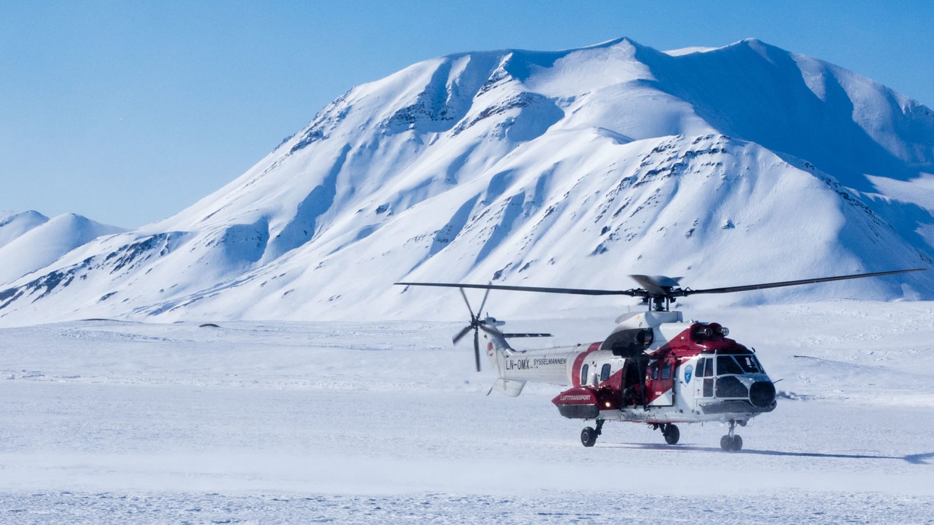 Weiß-roter Super Puma mit Kennung LN-OMX und Schriftzug Sysselmannen steht auf schneebedeckter Ebene vor verschneitem Berg