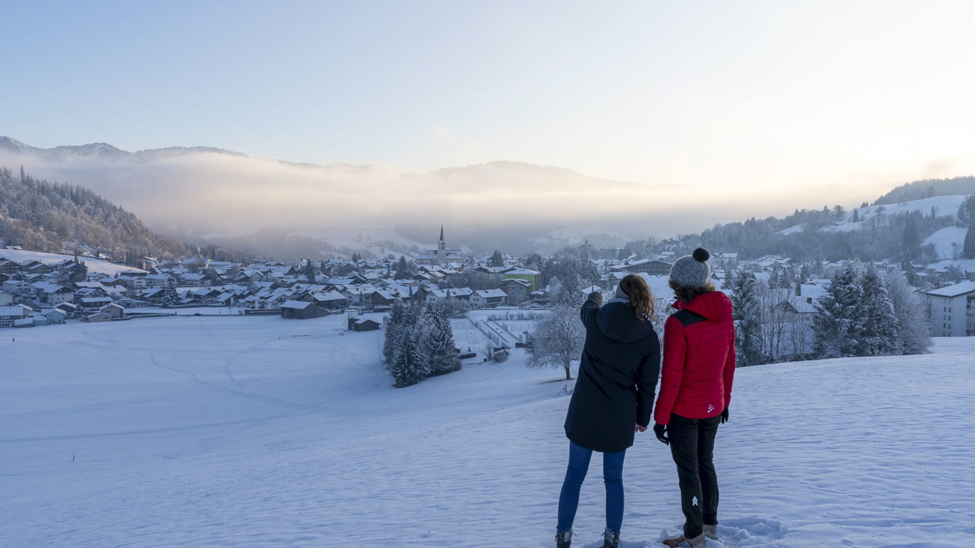Zwei Winterwanderer stehen auf verschneitem Hang und blicken auf die Ortschaft Oberstaufen im Morgenlicht.