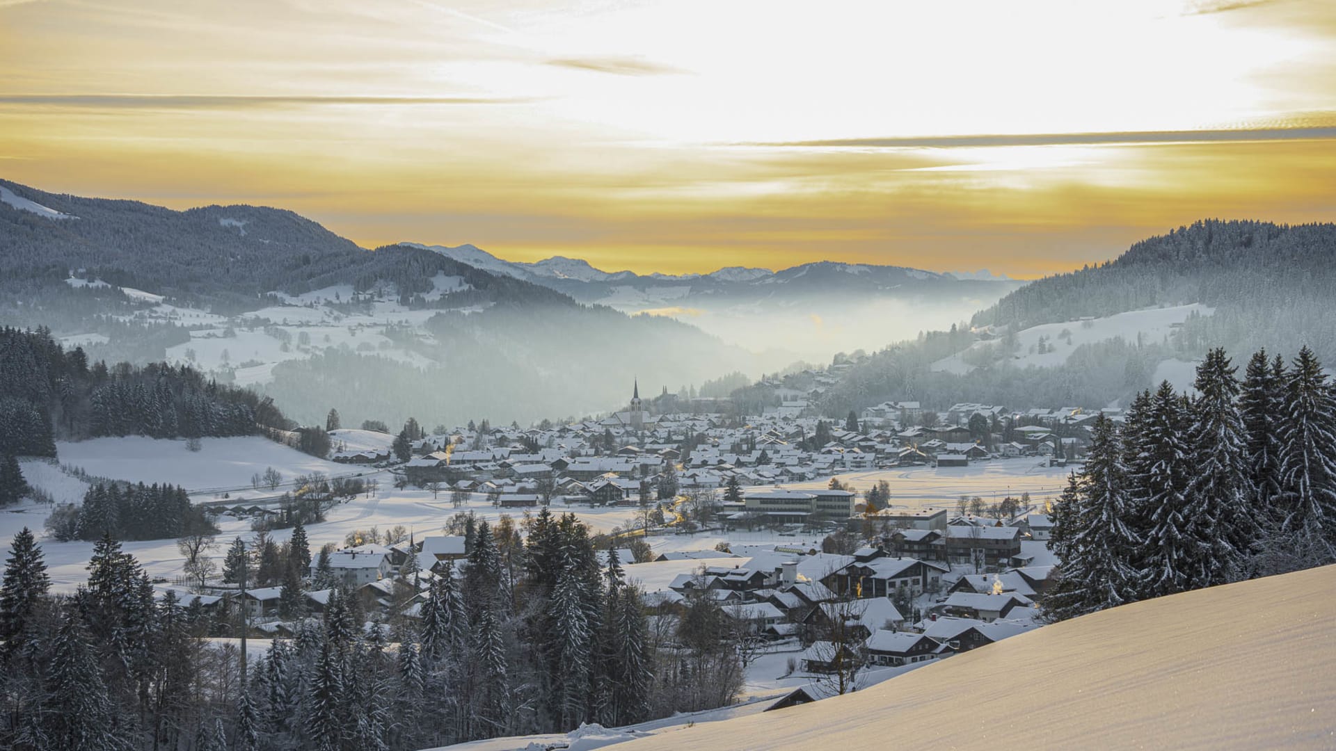 Winterwanderung bei Oberstaufen: Blick auf den verschneiten Ort mit Kirche und Alpenpanorama im Morgen- oder Abendlicht.