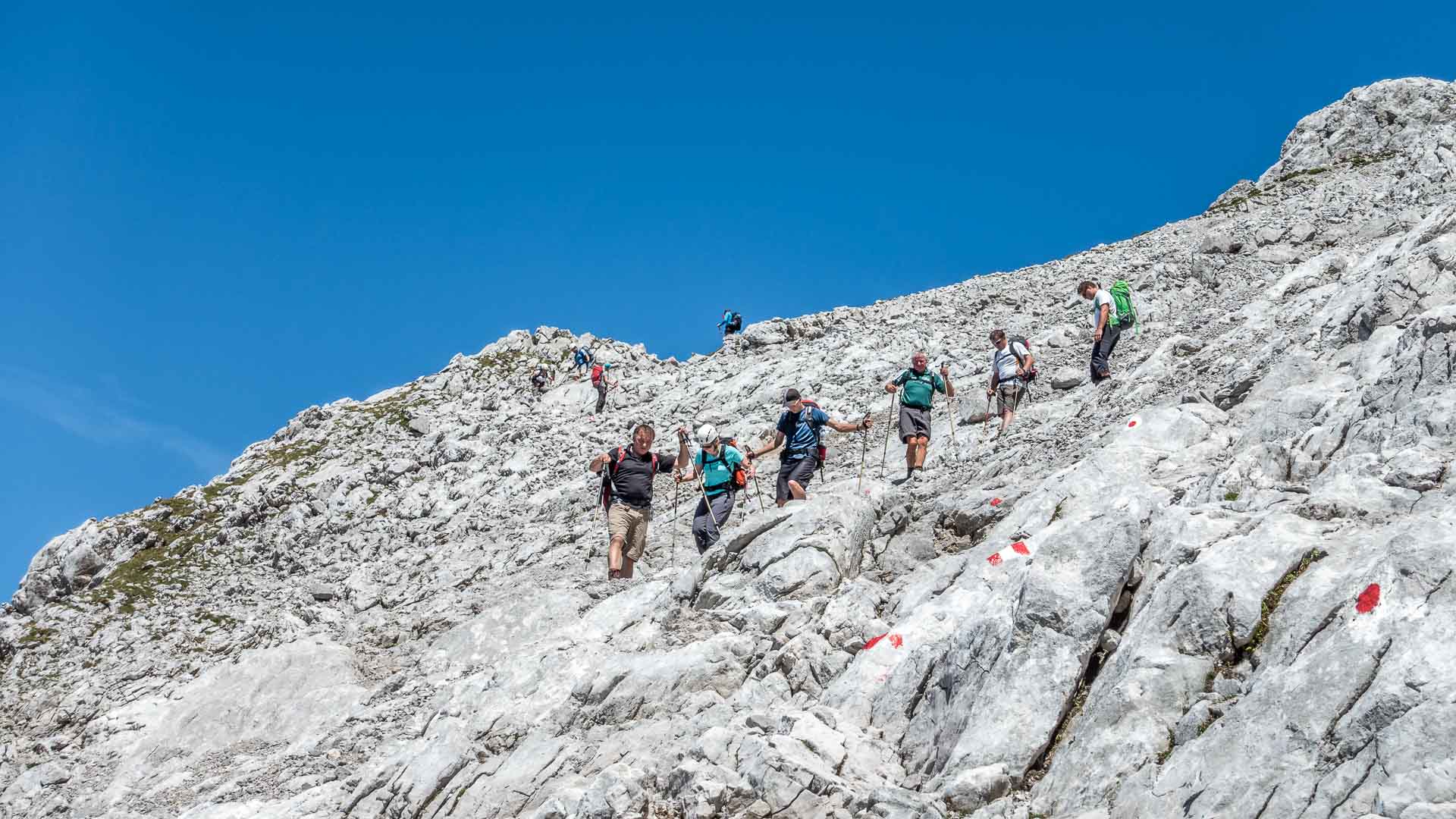 Acht Bergwanderer steigen über zerklüfteten Kalkfels am Watzmann ab, rote Wegmarkierungen am Fels sichtbar.