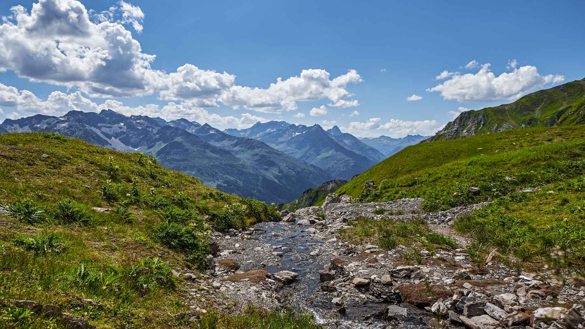 Bergwanderung im Lechtal: steiniger Bach im Vordergrund, grüne Almhänge und Bergkette mit Schneefeldern im Hintergrund.