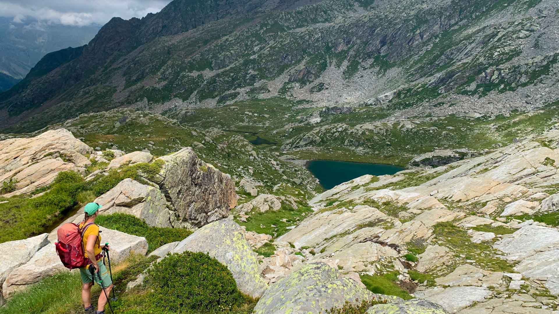 Bergwanderer mit rotem Rucksack und Wanderstock blickt auf türkisfarbenen Bergsee im Hochgebirgskessel des Piemont.