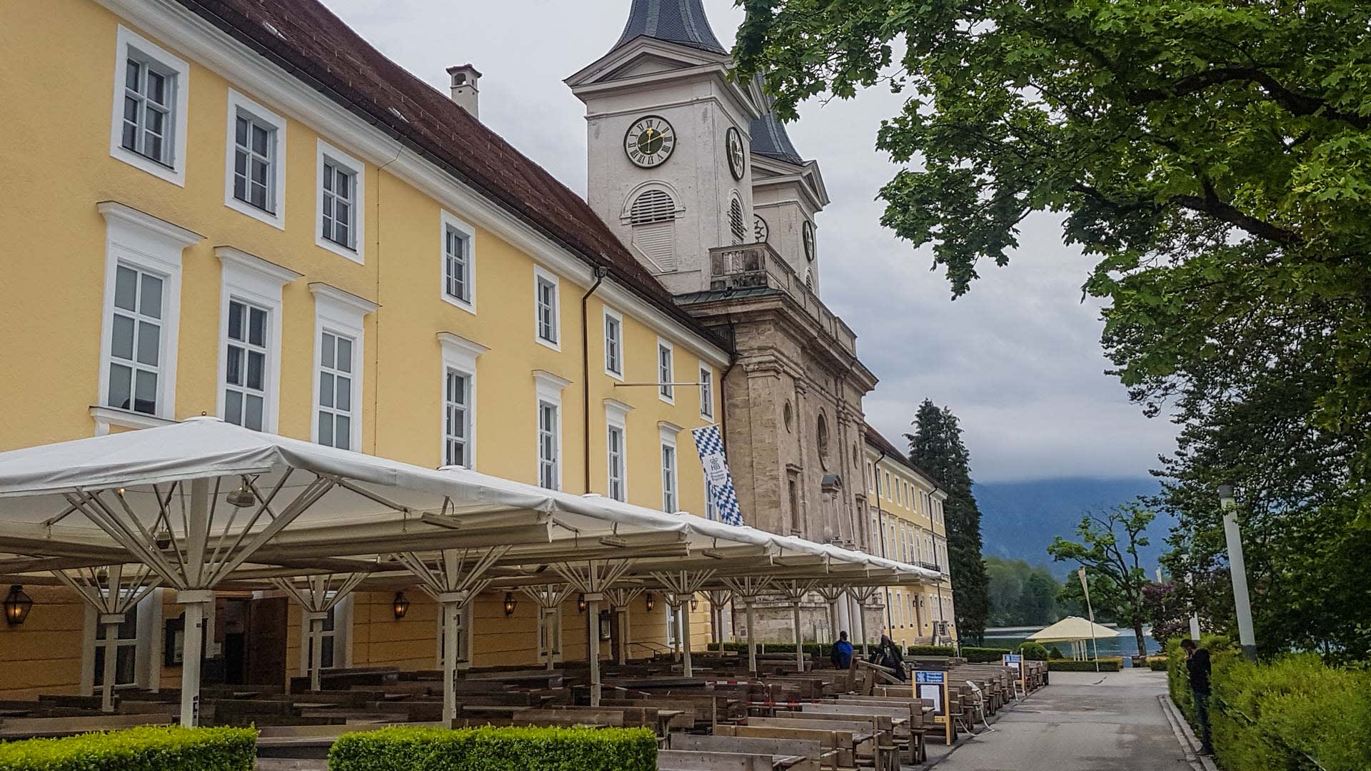 Mehrgeschossiges Gasthaus mit gelbem Fassadenputz, Kirchturm und überdachtem Biergarten am Tegernsee – Einkehrziel