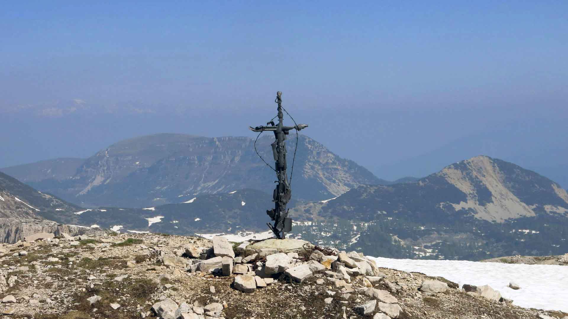 Bergwanderung auf den Dente Austriaco: metallenes Gipfelkreuz auf Steinmann mit Bergpanorama der Vizentinischen Alpen im Sommer.