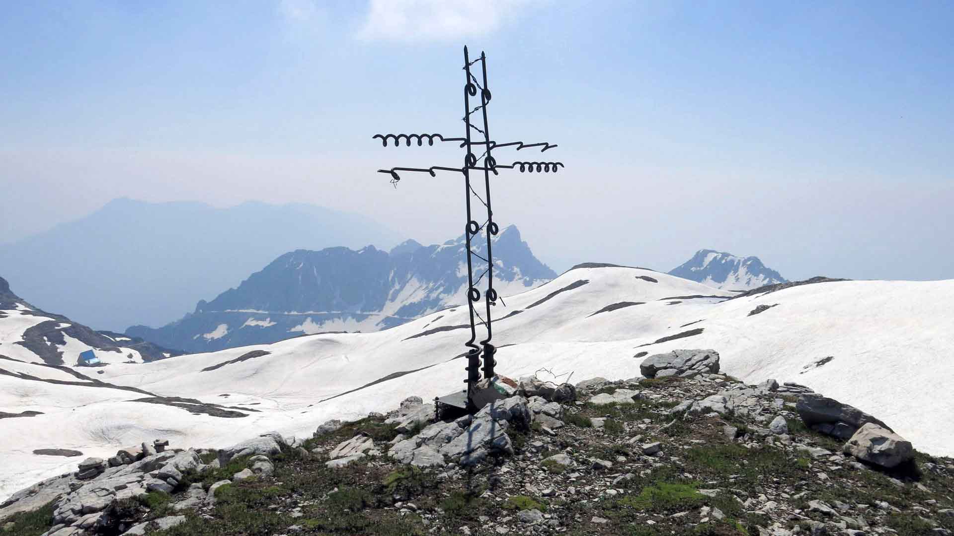 Bergwanderung auf den Dente Italiano: schmiedeeisernes Gipfelkreuz auf felsigem Gipfel mit Bergkämmen im Hintergrund.