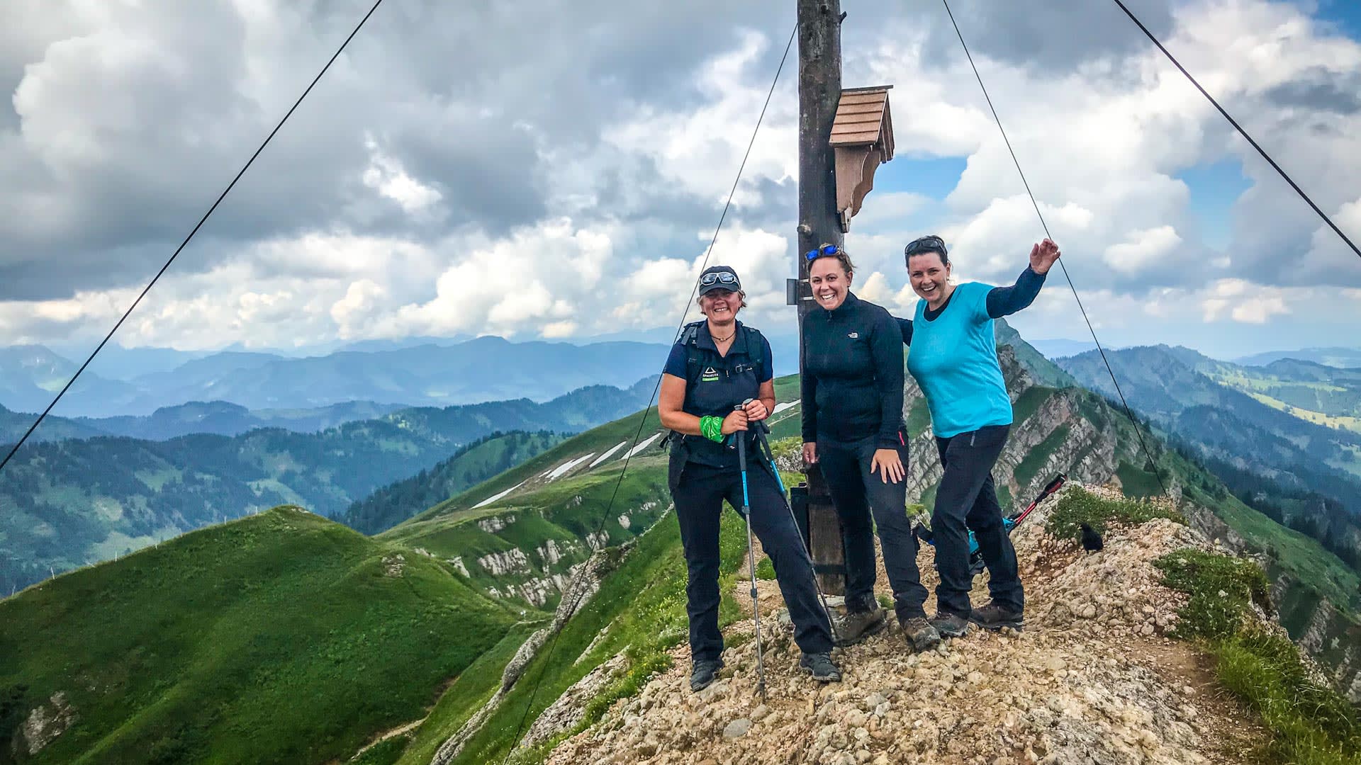 Drei Bergwandererinnen am Gipfelkreuz des Rindalphorns auf der Nagelfluhkette mit Blick auf grüne Bergkämme im Allgäu.