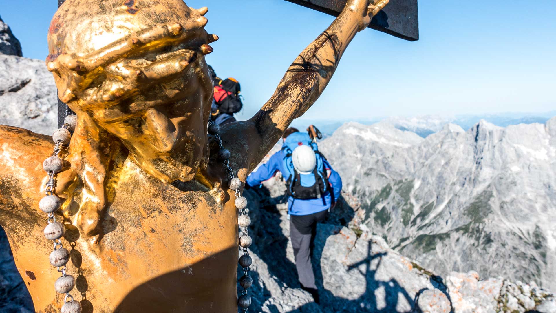Bergwanderung am Watzmann Hocheck: vergoldetes Gipfelkreuz mit Christusfigur und Rosenkranz, zwei Bergwanderer im Hintergrund.