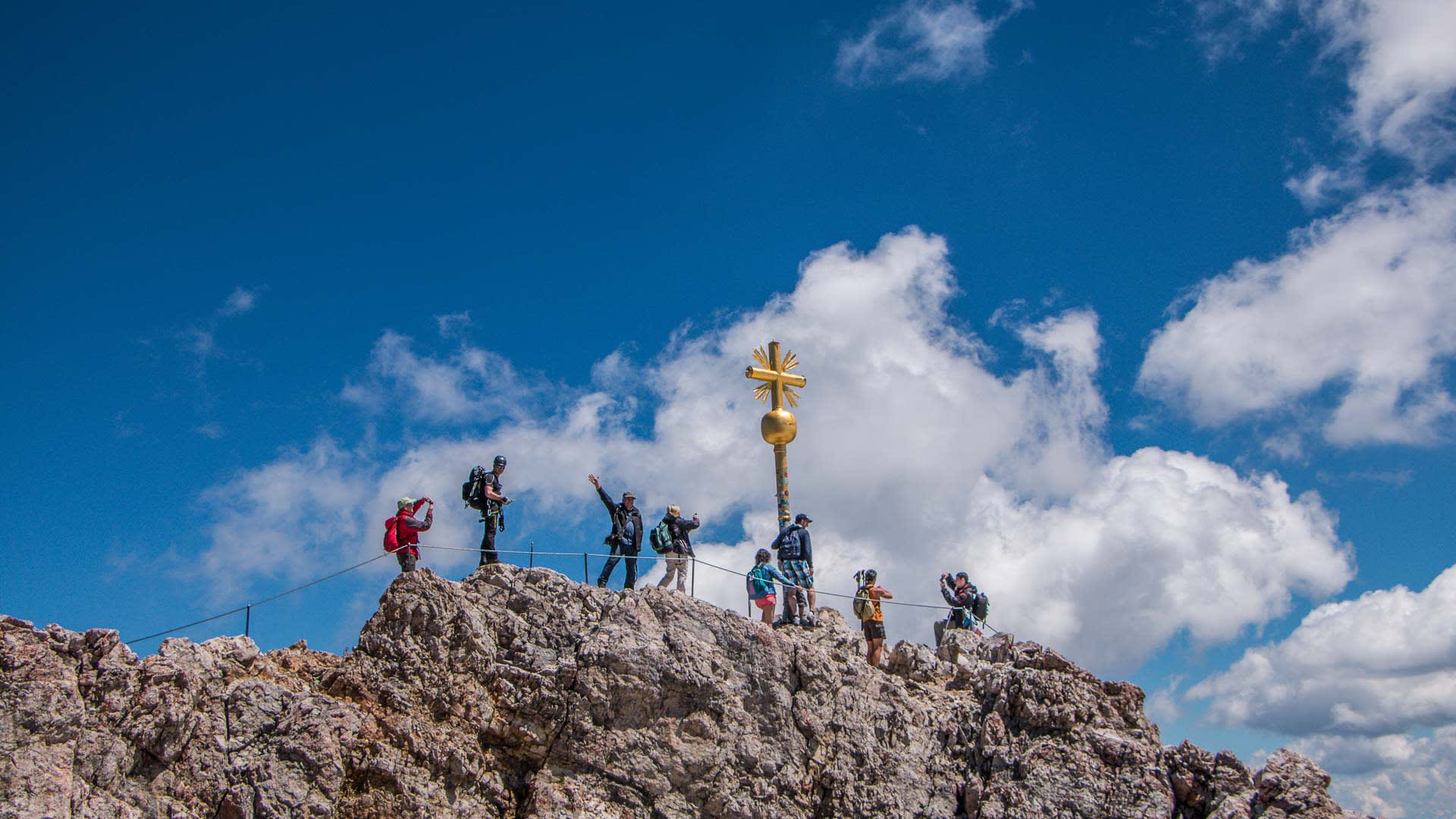 Bergwanderung auf die Zugspitze: sieben Bergwanderer am goldenen Gipfelkreuz auf dem felsigen Grat.