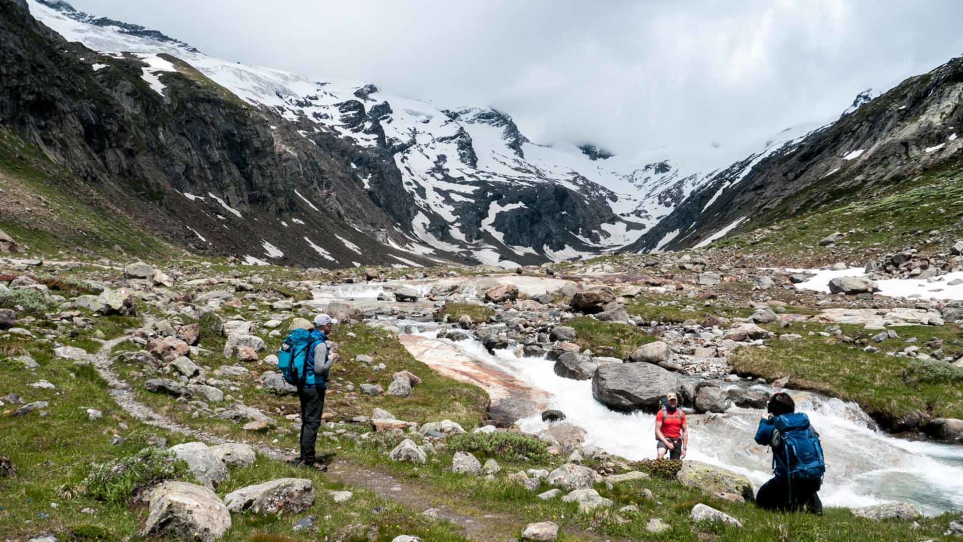 Drei Bergwanderer rasten an einem Gletscherbach in einem hochalpinen Tal der Hohen Tauern mit Schneefeldern im Hintergrund.