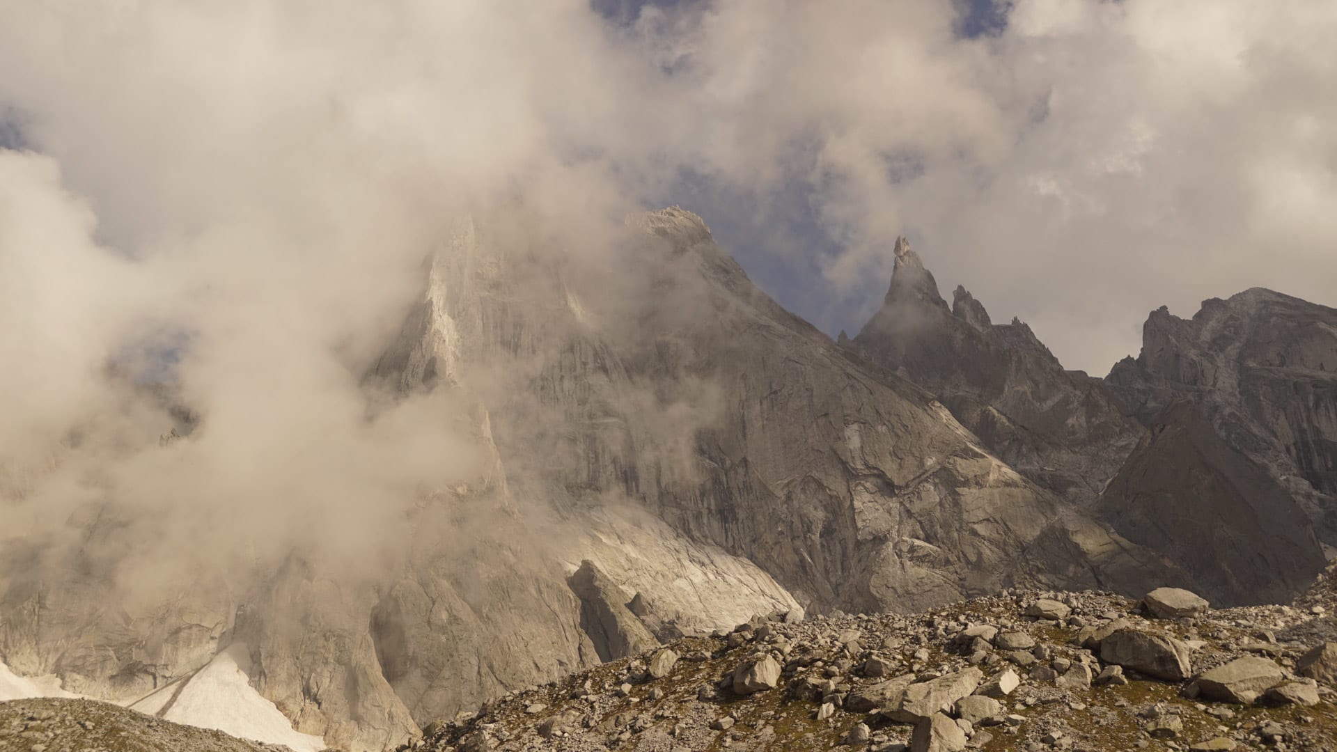 Massive Granitwand mit spitzem Gipfel und Wolkenschwaden über Geröllplateau im Bergell im Sommer.