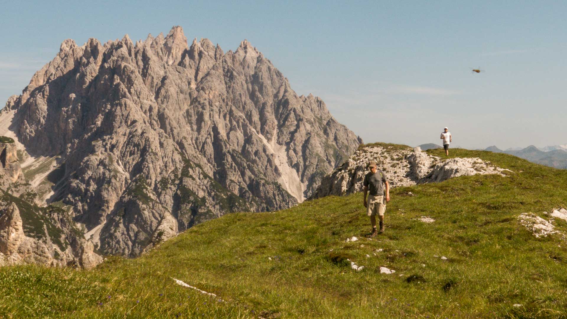 Zwei Bergwanderer steigen auf dem Graskamm im Wilden Kaiser auf, hinter ihnen eine Felswand mit gezackten Kalksteingipfeln.