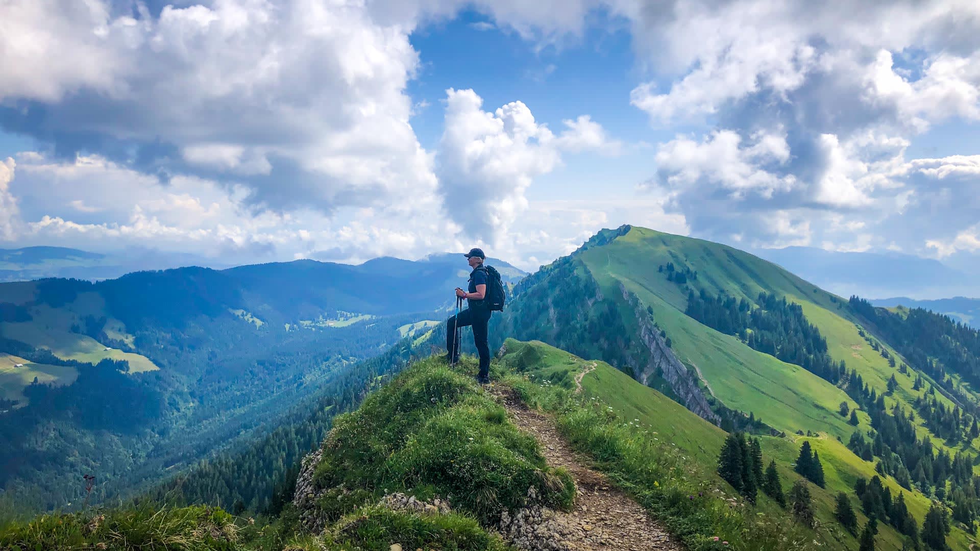 Bergwanderer steht auf Gratpunkt in den Allgäuer Voralpen, Wanderpfad führt zum grasbewachsenen Nachbargipfel.