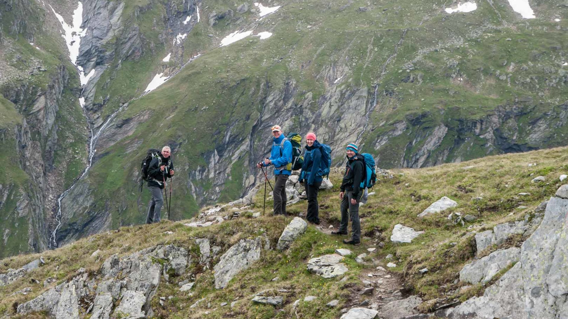 Vier Personen bei der Bergwanderung in den Hohen Tauern auf steinigem Pfad am Berghang mit Wasserfall und Schneefeldern.