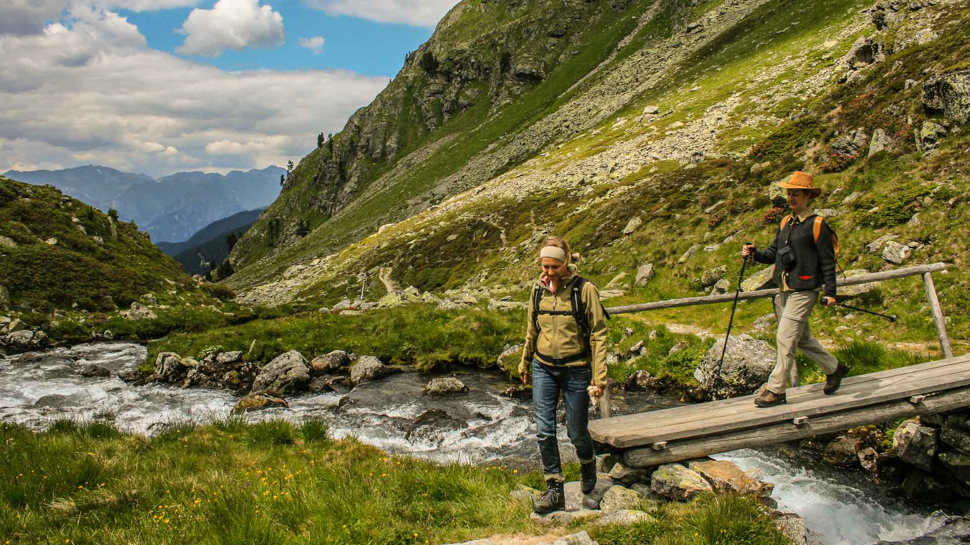 Zwei Bergwanderer überqueren eine Holzbrücke über einen Gebirgsbach in den Ammergauer Alpen im Sommer.