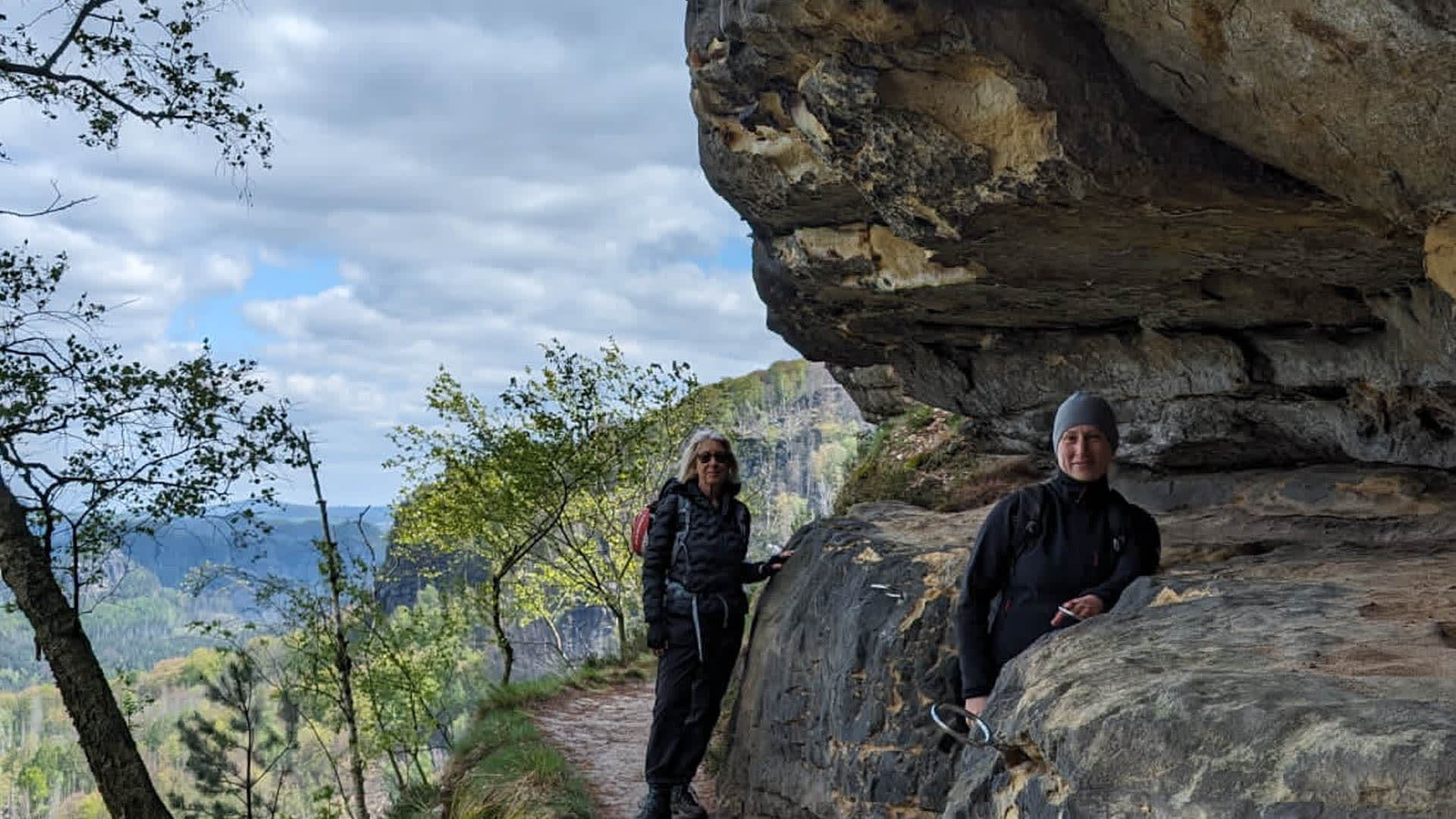 Zwei Wanderer pausieren bei der Bergwanderung in der Sächsischen Schweiz an einer markanten Sandsteinfelsenwand mit Talblick.