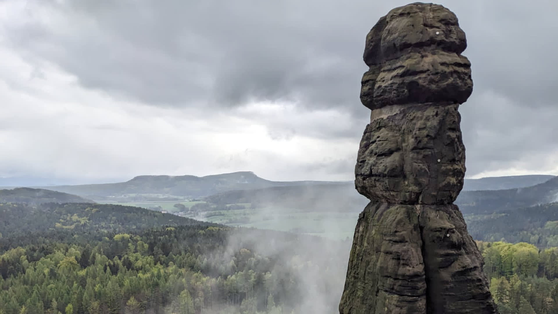 Bergwanderung in der Sächsischen Schweiz: schlanker Sandsteinfelsen mit Kletterbolzen vor bewaldeter Tallandschaft.