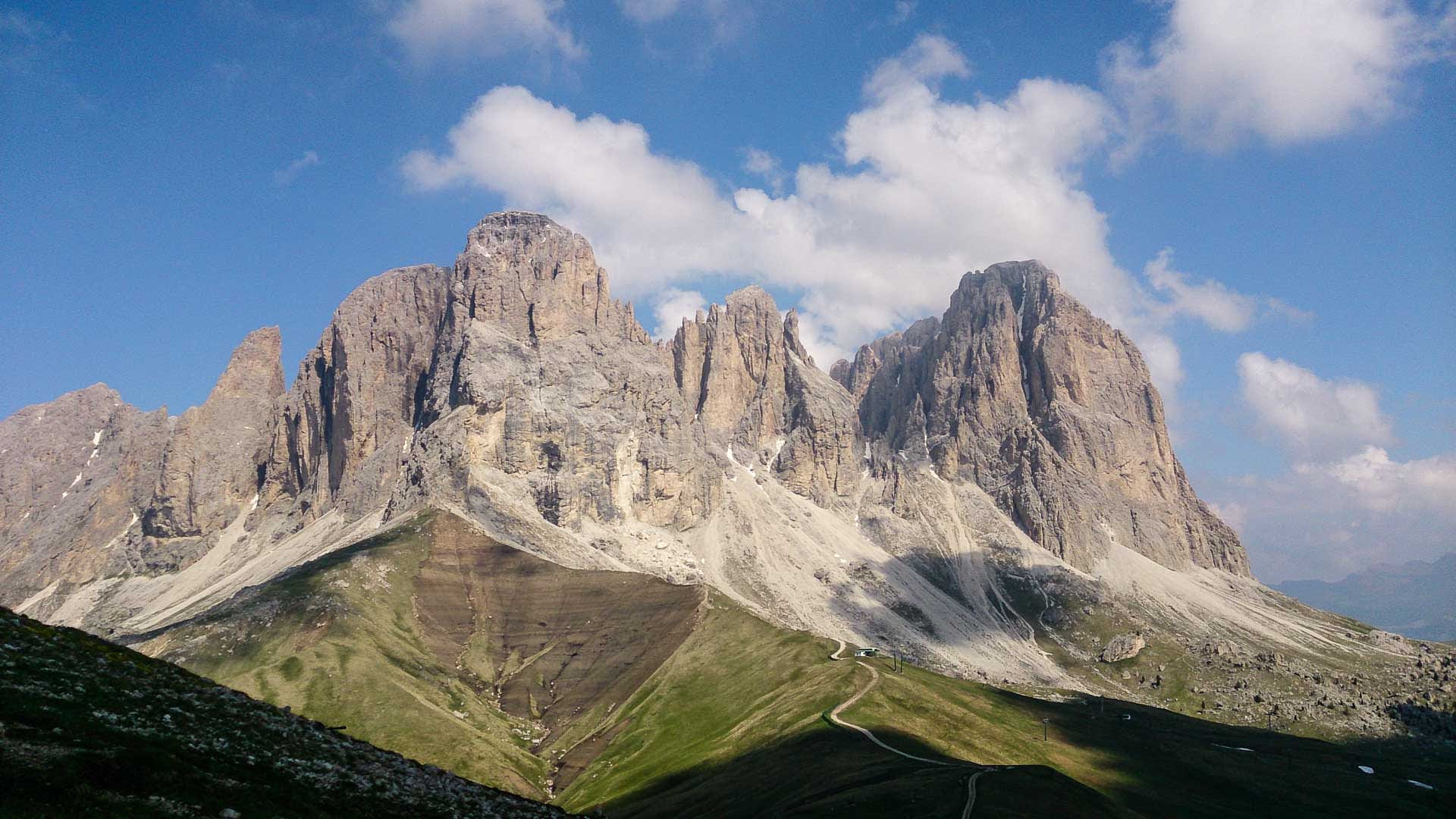 Bergwanderung am Sella Pass: Kalkfels-Massiv mit senkrechten Türmen, Schrofenhängen, Almwiesen und geschwungenem Fahrweg im Sommer.