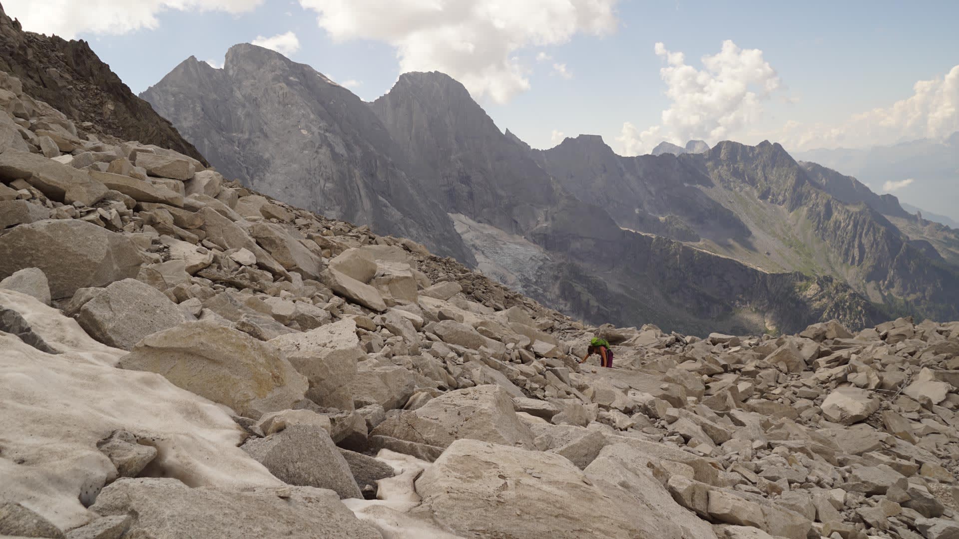 Bergwanderer mit grünem Rucksack überquert einen Geröllhang beim Sentiero Roma im Bergell, im Hintergrund markante Felsgipfel