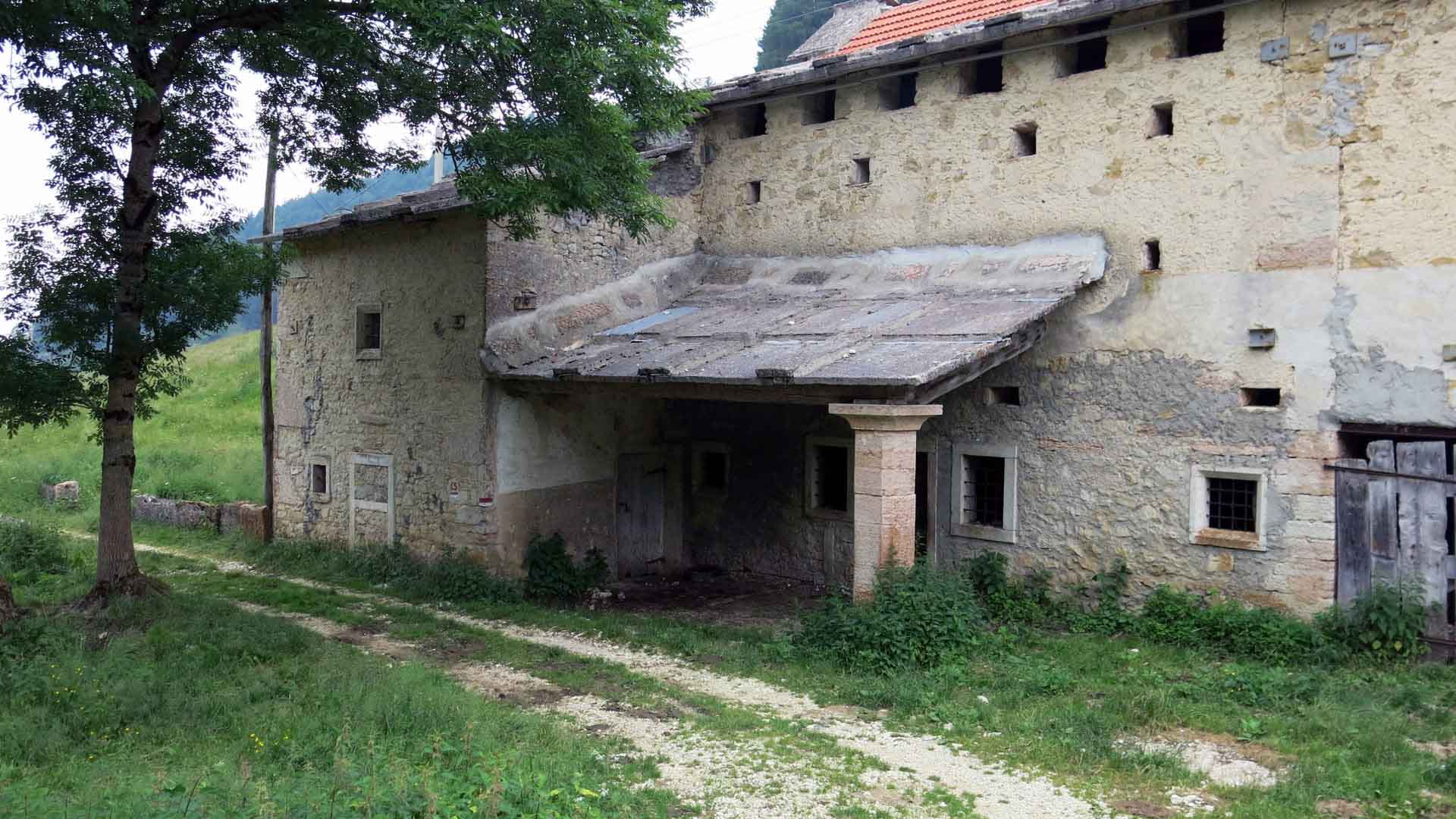 Bergwanderung bei Erbezzo: verwittertes mehrstöckiges Bruchsteingebäude mit überdachtem Eingang in ländlicher Sommerlandschaft.
