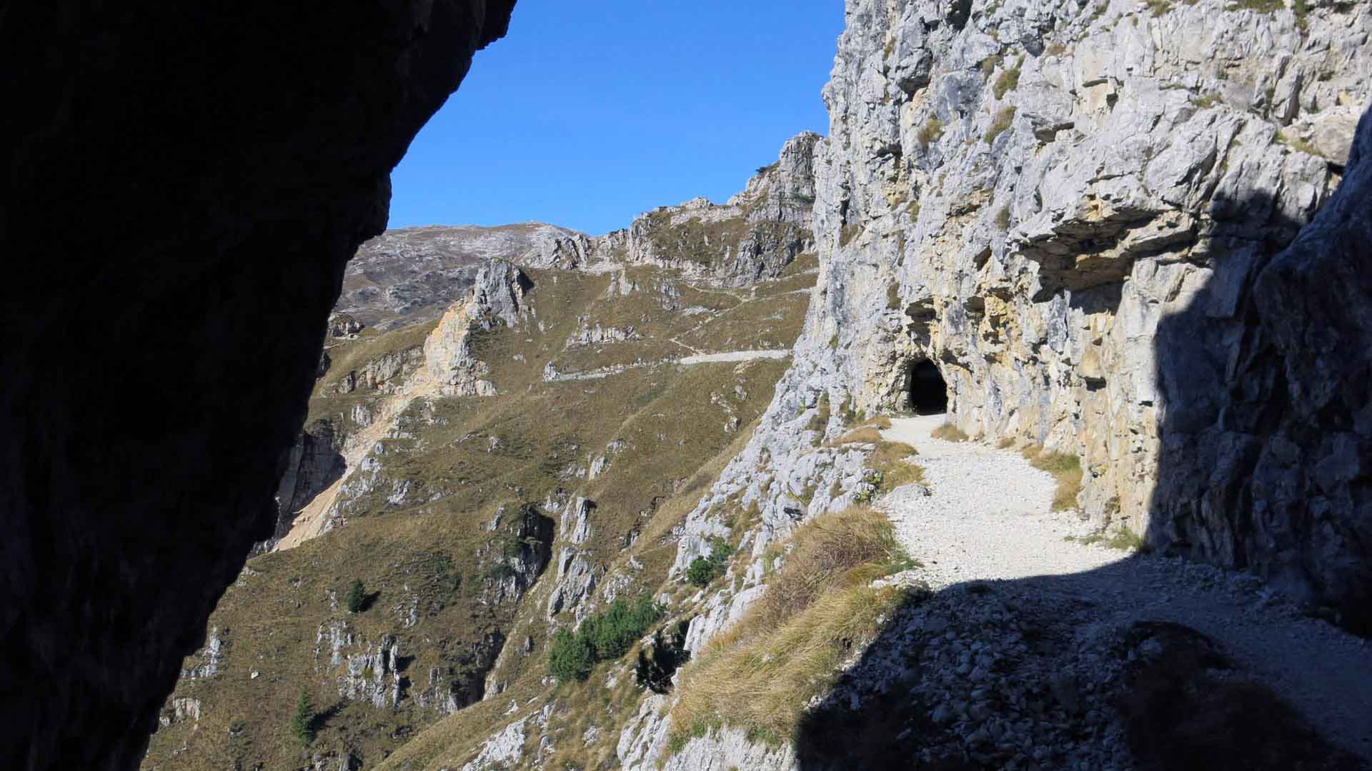Bergwanderung auf der Strada della 52 – Schotterweg entlang der Kalkfelswand mit Blick auf einen Tunneleingang.