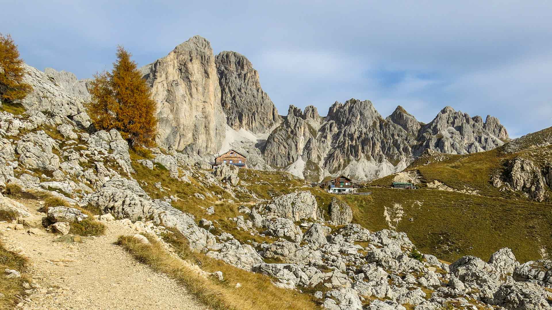 Bergwanderung zu den Vajolet Türmen im Rosengarten: Schotterweg mit Berghütte vor senkrecht aufragenden Kalksteintürmen im Herbst.