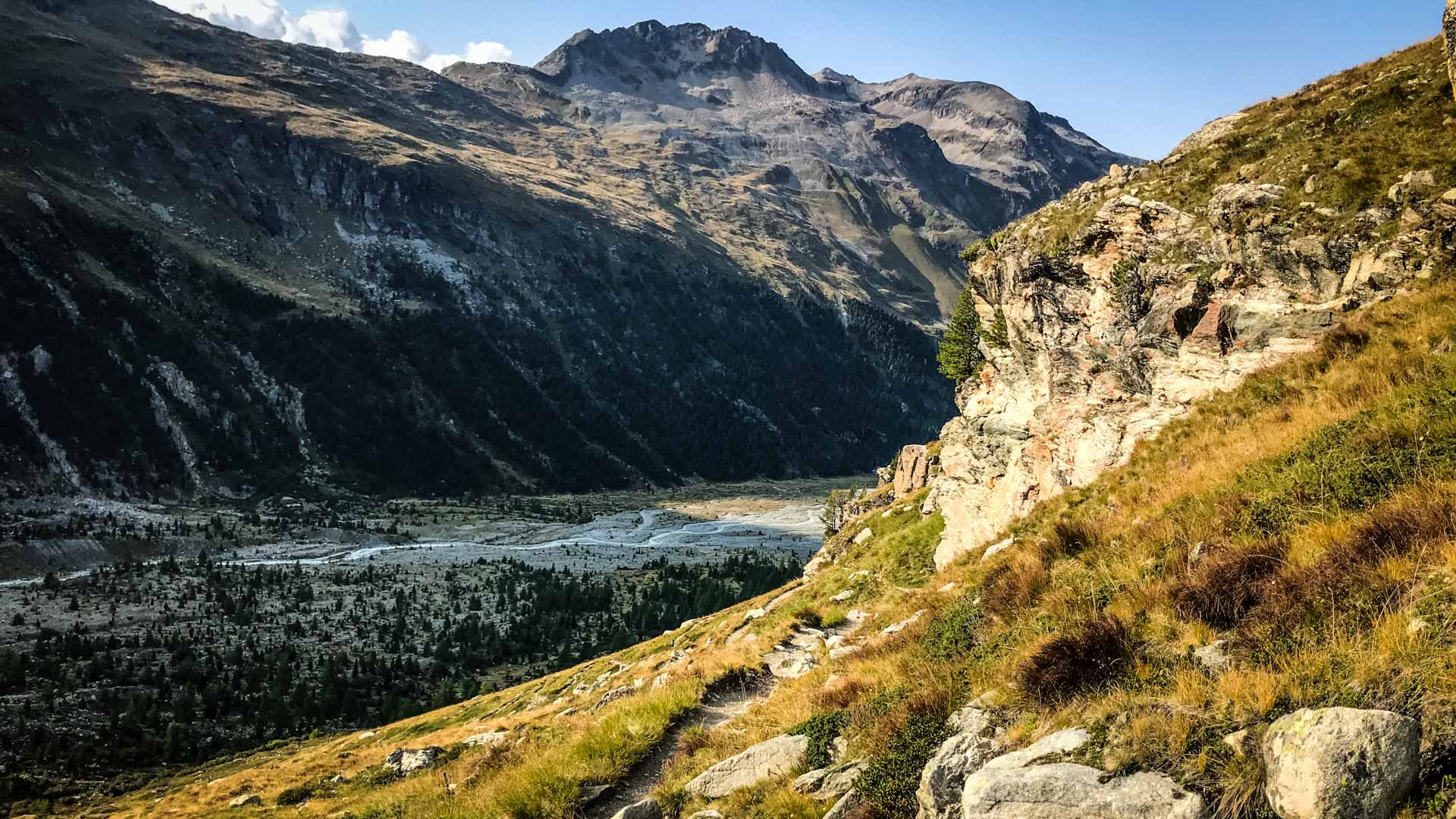 Bergwanderung im Val Roseg: Weites Alpental mit verzweigtem Flussbett, Nadelwald und felsigem Gipfel im Hintergrund.