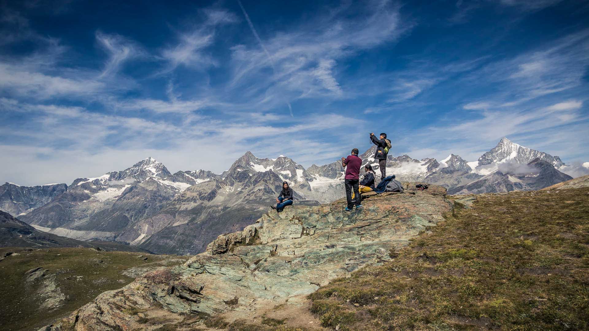 Vier Personen bei der Bergwanderung auf felsigem Aussichtspunkt mit Blick auf Gletscher und Viertausender der Schweizer Alpen.