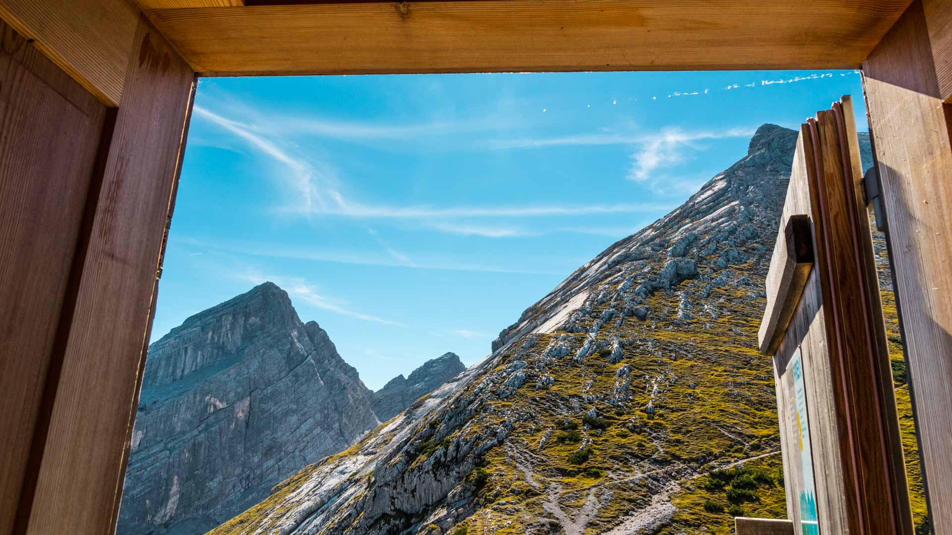 Bergwanderung am Watzmann: Blick durch Holzrahmen des Watzmannhauses auf Felsweg und Gipfelturm im Sommer.
