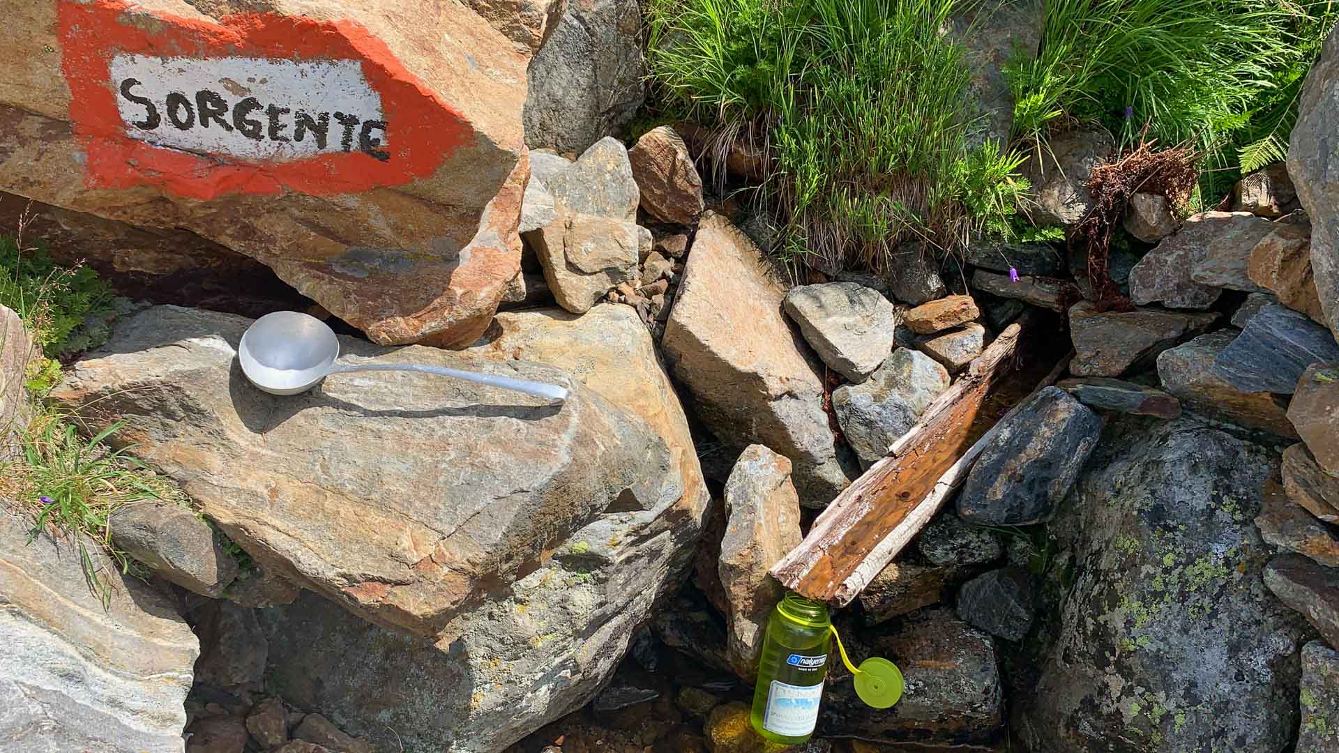 Bergwanderung GTA Piemont: roter Wegpfeil mit Aufschrift SORGENTE an Fels, Schöpfkelle und Trinkflasche.