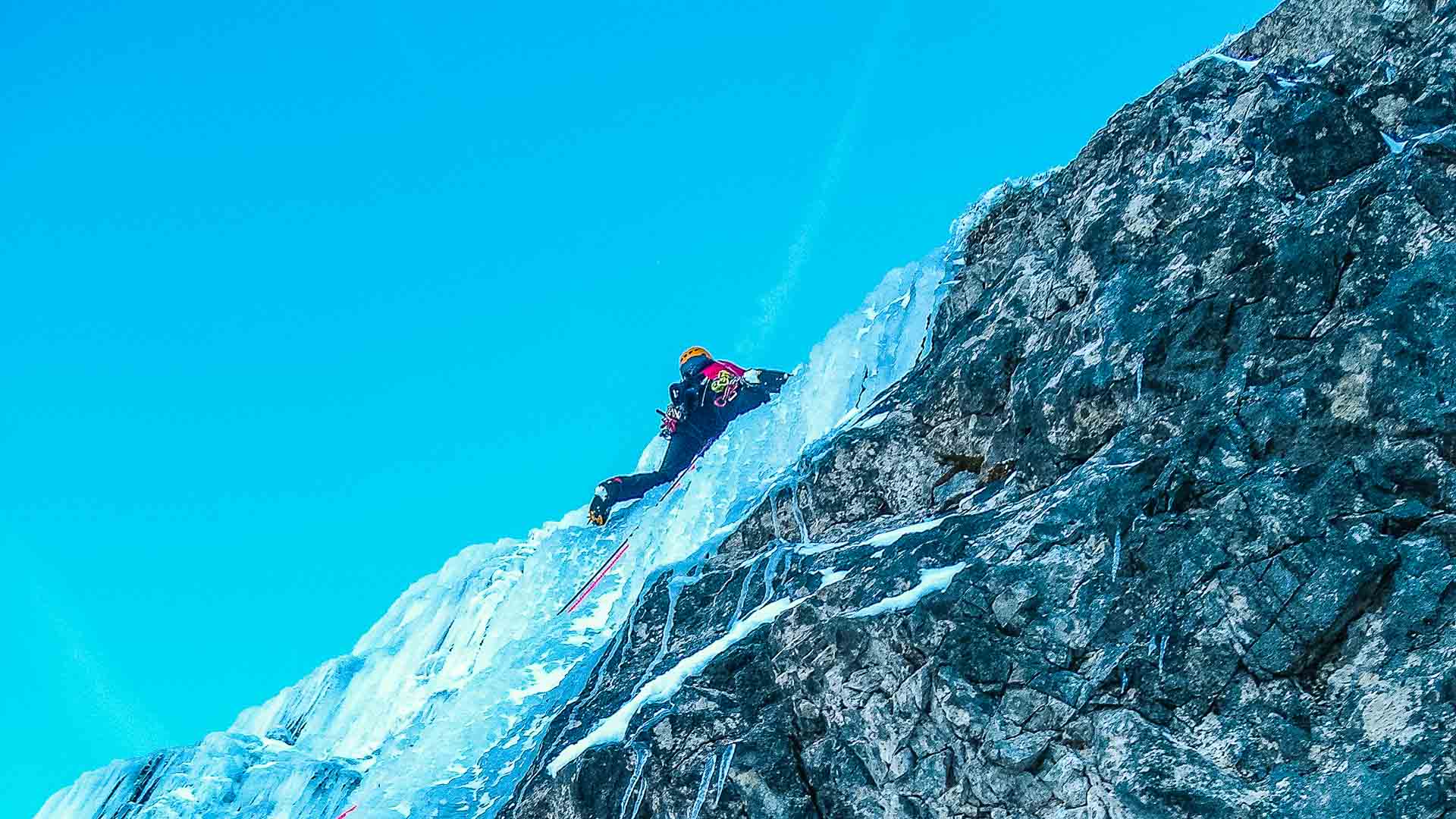Eiskletterer mit Helm und Steigeisen klettert an schmaler Eisformation vor türkisblauem Himmel im Allgäu.