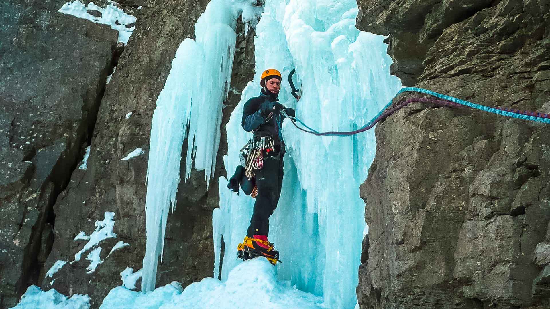 Eiskletterer steht mit Helm, Steigeisen und Eispickel vor blauem Eiswasserfall beim Eiskletterfestival in Kandersteg.