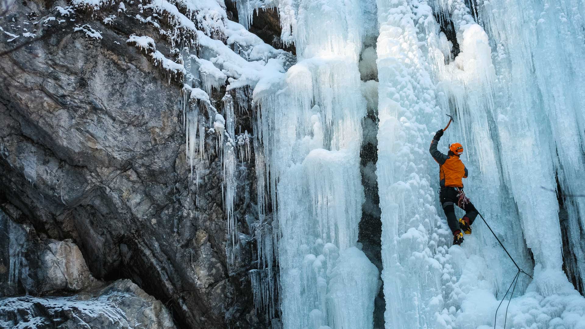 Eiskletterer in oranger Jacke und Helm schlägt Pickel in eine senkrechte Eiswand mit massiven Eissäulen in Bad Gastein.