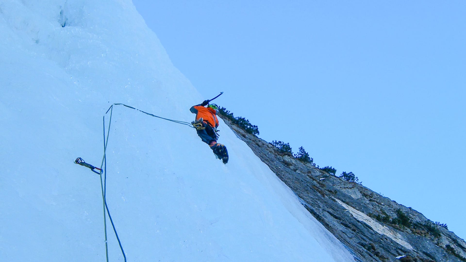 Eiskletterer in oranger Jacke und grünem Helm klettert eine Mehrseillängentour an blau-weißer Eiswand in Bad Gastein aufwärts.