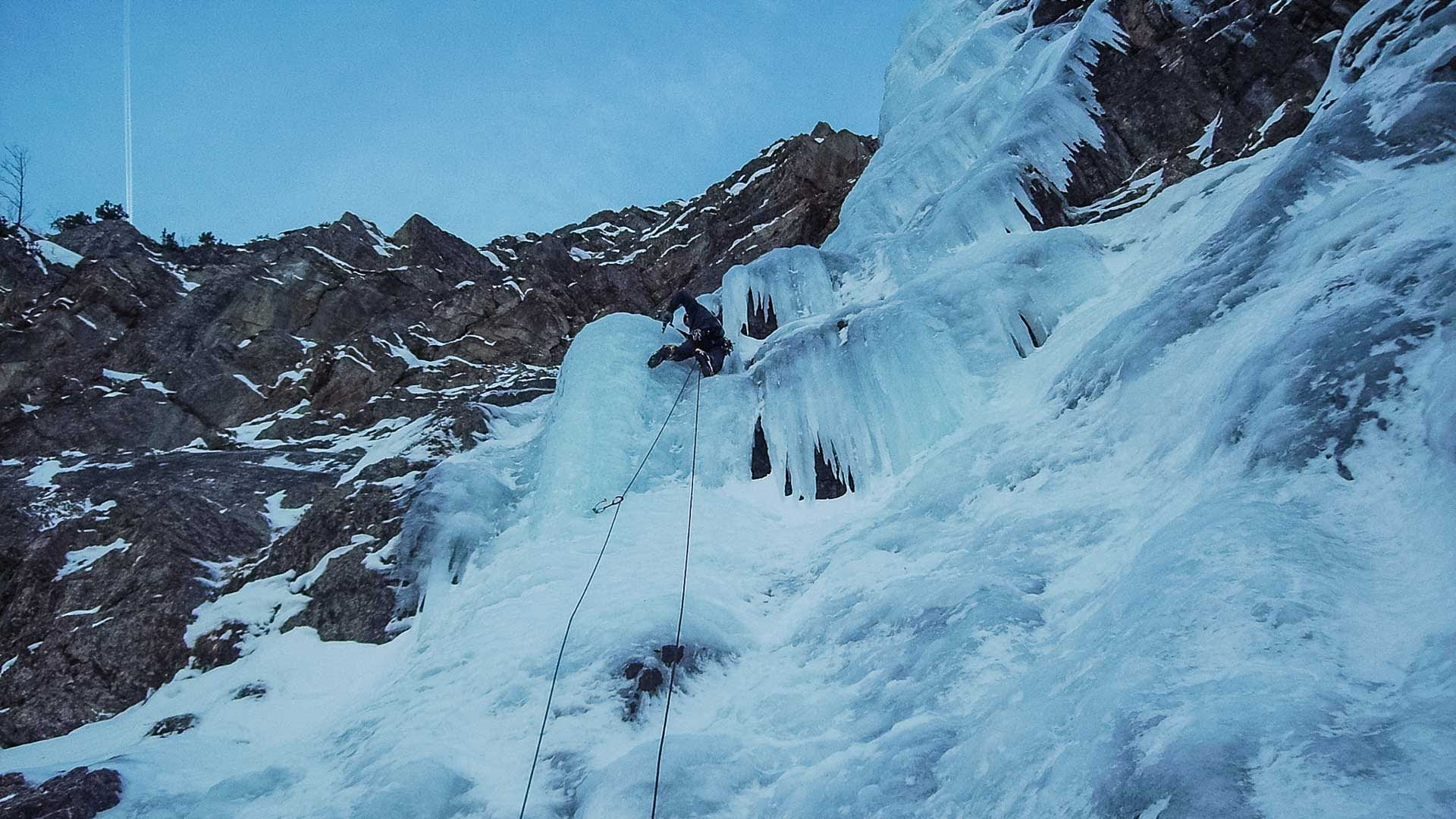 Eiskletterer steigt im Vorstieg eine steile blaue Eiswand in Bad Gastein auf, Seil und Zwischensicherung sichtbar.