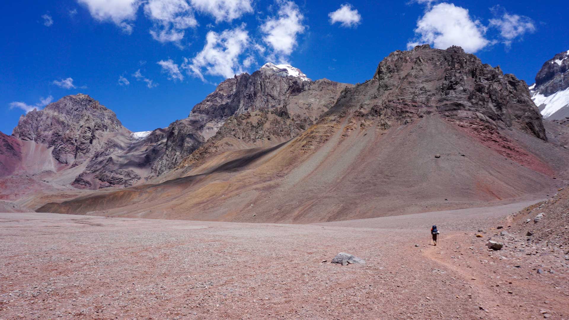 Person mit blauem Rucksack auf Schotterweg in vegetationsloser Anden-Hochgebirgslandschaft bei Aconcagua-Expedition