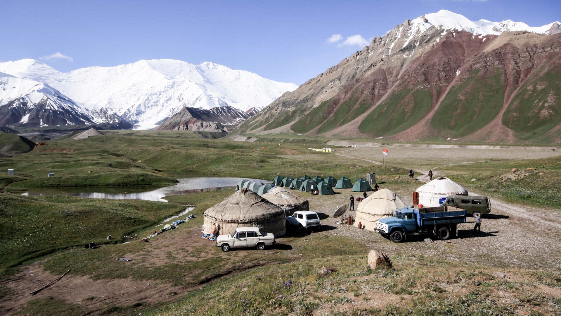Expedition Pik Lenin: Basislager Achik Tasch mit Jurten, Zelten und Fahrzeugen im Hochtal vor verschneiten Gipfeln.
