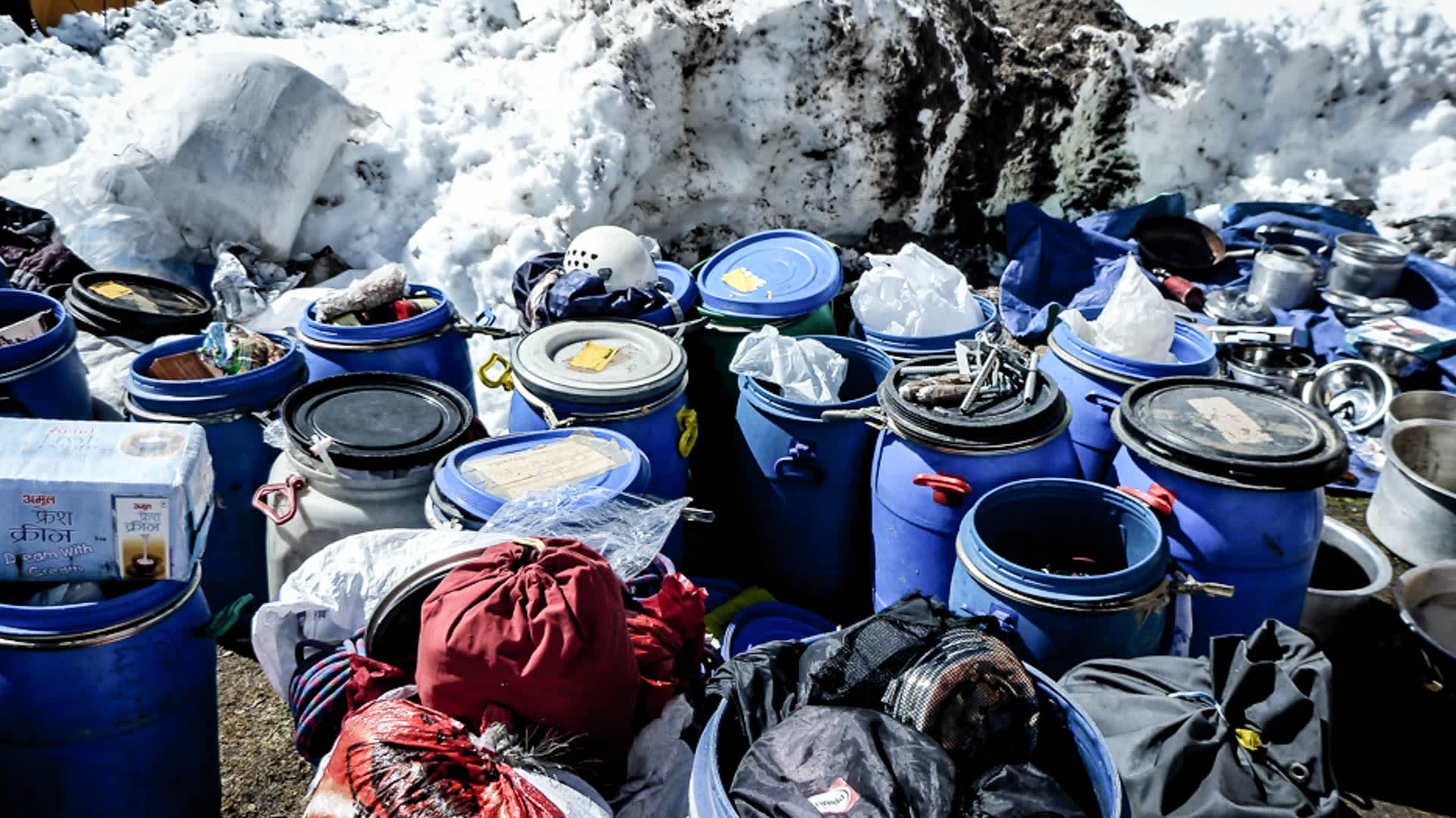 Expedition Ama Dablam: blaue Versorgungsfässer, Duffle-Bags und Ausrüstung im Basislager vor Schneewand.