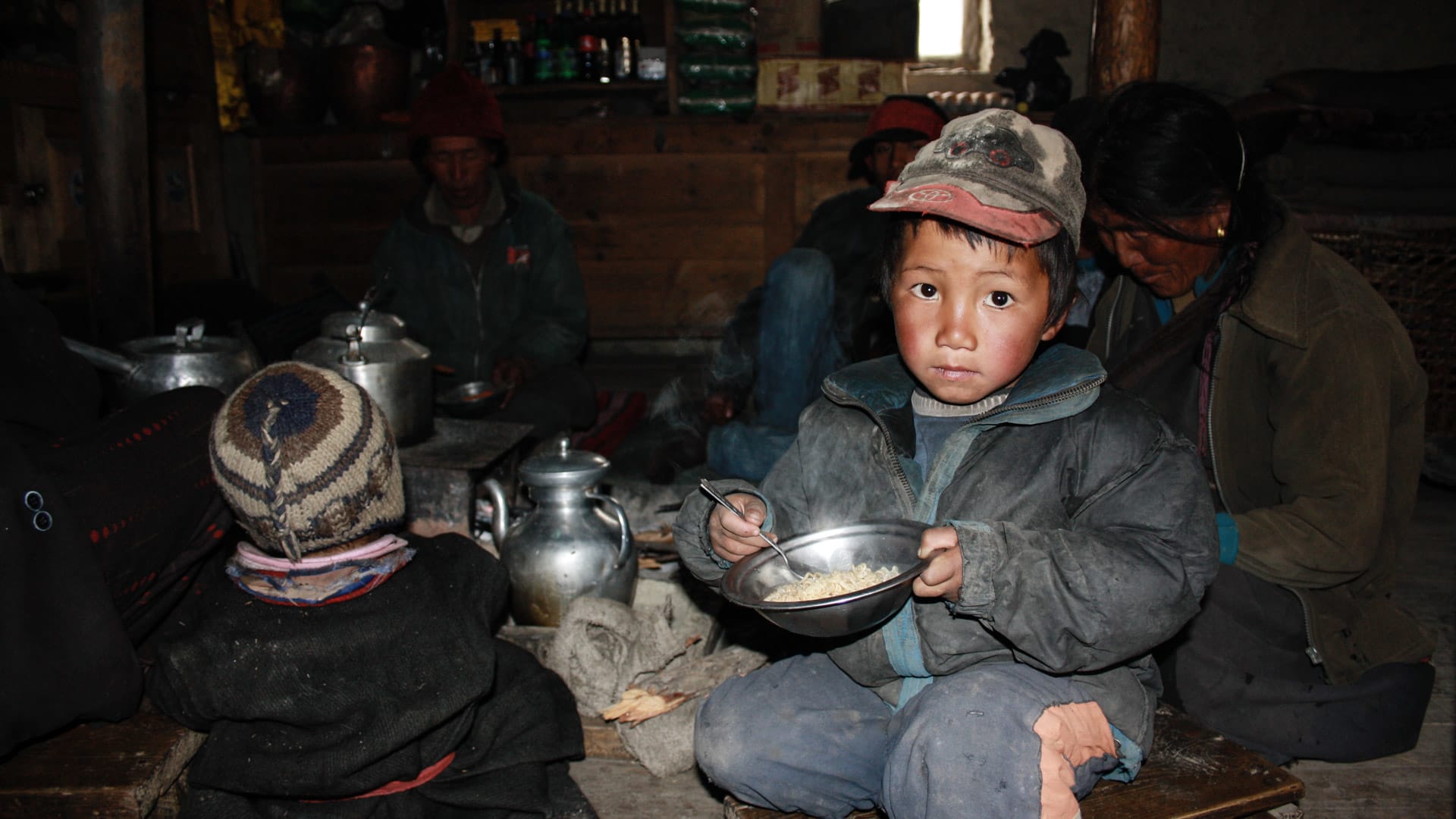 Expedition Himlung Himal: Kind mit Metallschüssel und drei weitere Personen beim Buttertee in einem rustikalen Teehaus in Tibet.