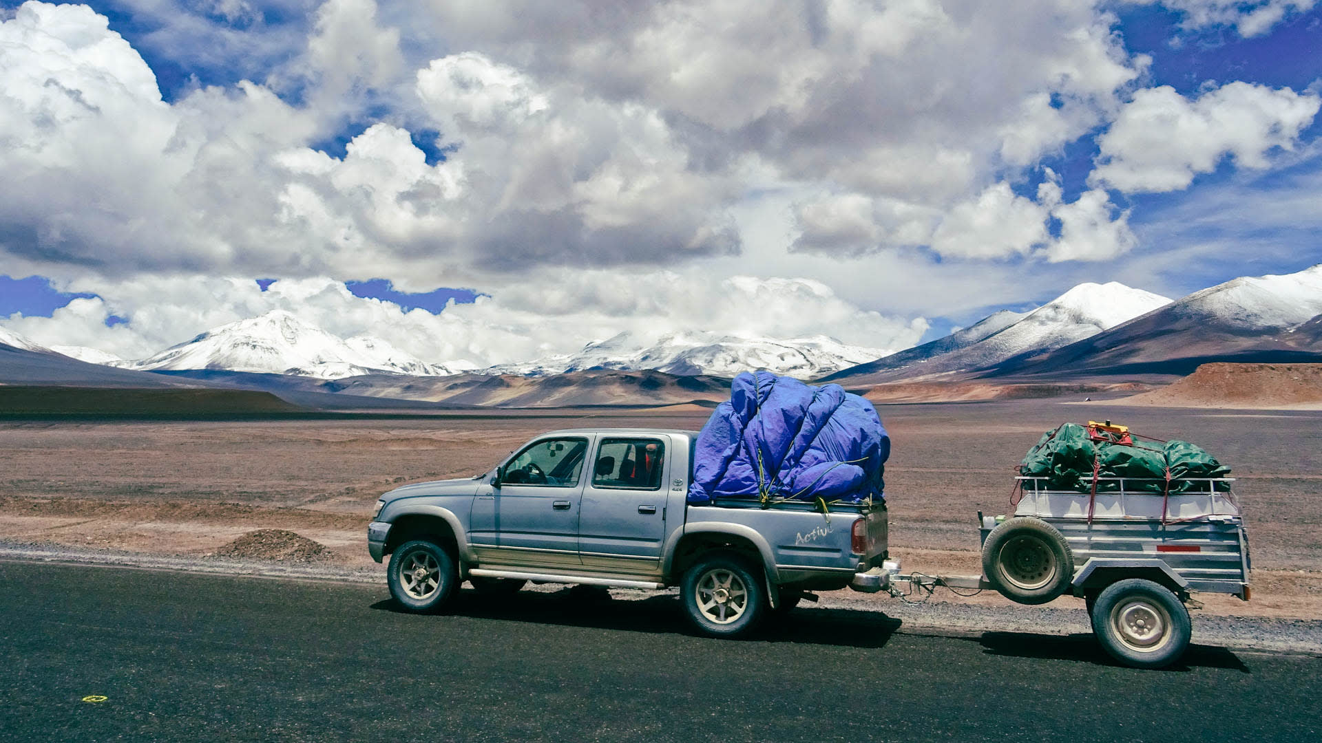 Pickup-Truck mit Anhänger und Expeditionsausrüstung auf asphaltierter Straße im Atacama-Hochplateau, schneebedeckte Gipfel