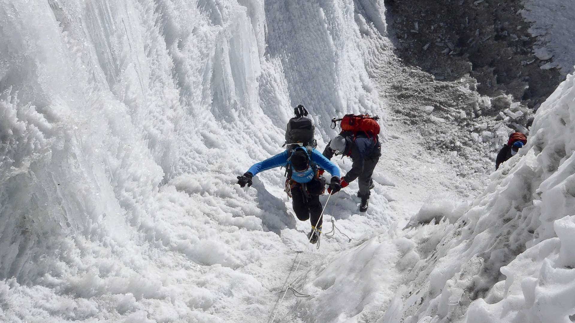 Drei Höhenbergsteiger mit Klettergurt und Steigeisen steigen an einem Fixseil eine Steilwand am Amphu-Lapsa-Pass auf.