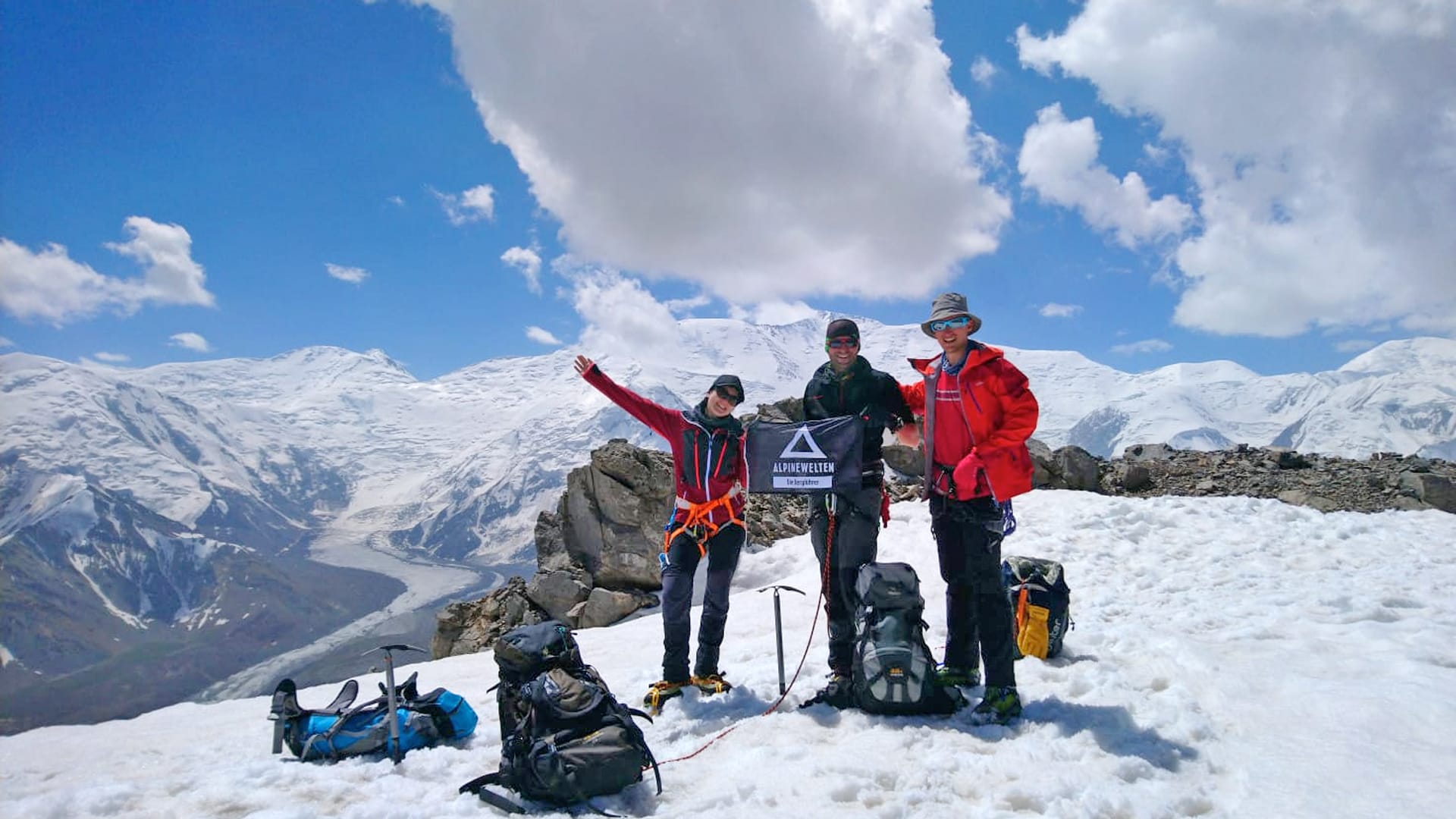 Expedition Pik Lenin: Drei Höhenbergsteiger mit Alpinewelten-Banner, Eispickel und Seil auf schneebedecktem Gipfelplateau.