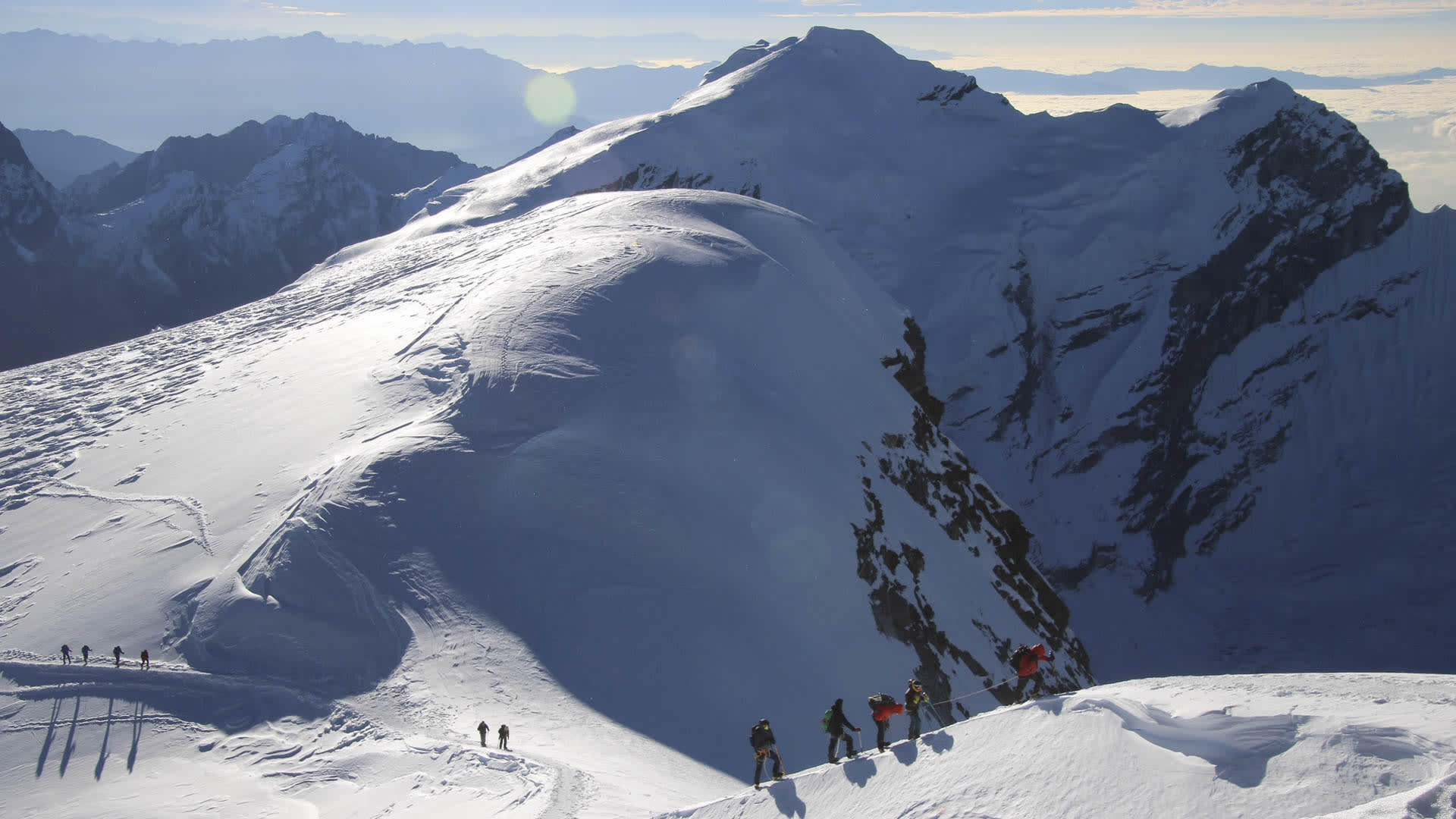 Expedition Mera Peak: Zwei Seilschaftsgruppen von Höhenbergsteigern bewegen sich bei Gegenlicht über den Schneegrat zum Gipfel.