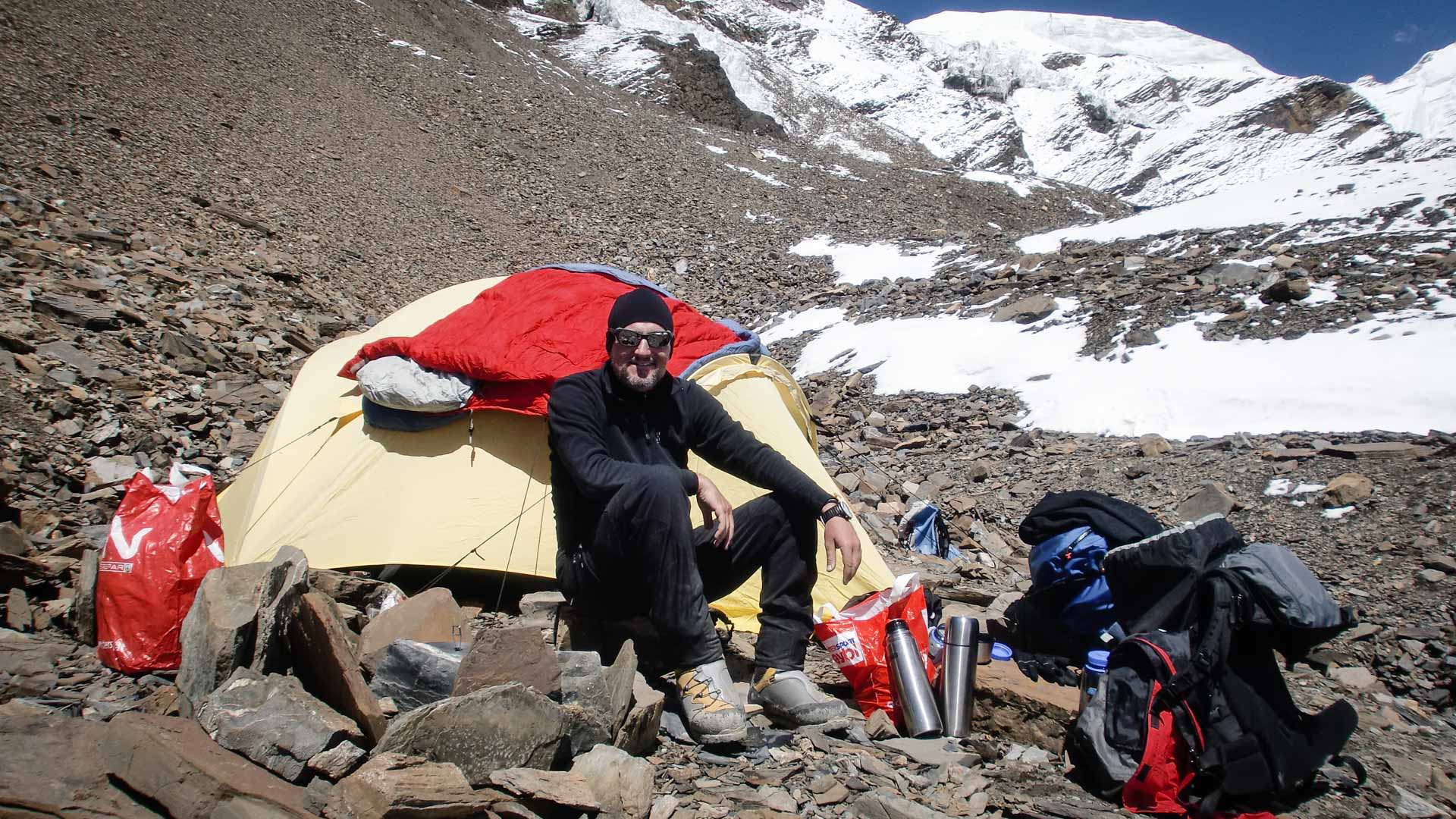 Höhenbergsteiger rastet vor einem gelb-roten Kuppelzelt in Lager I der Himlung Himal Expedition auf felsigem Untergrund.