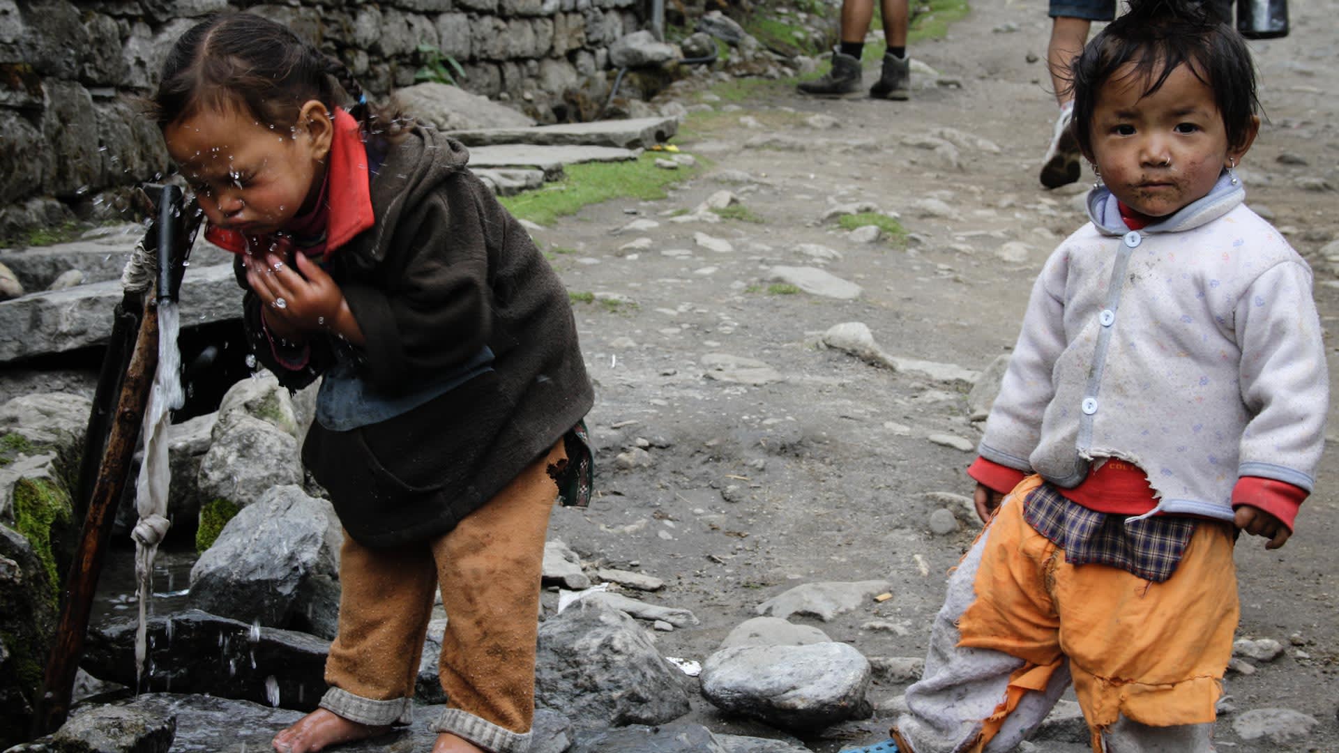 Zwei nepalesische Kleinkinder an einem Wasserauslauf auf einem Steinweg in einer Bergsiedlung entlang der Route zur Himlung Himal