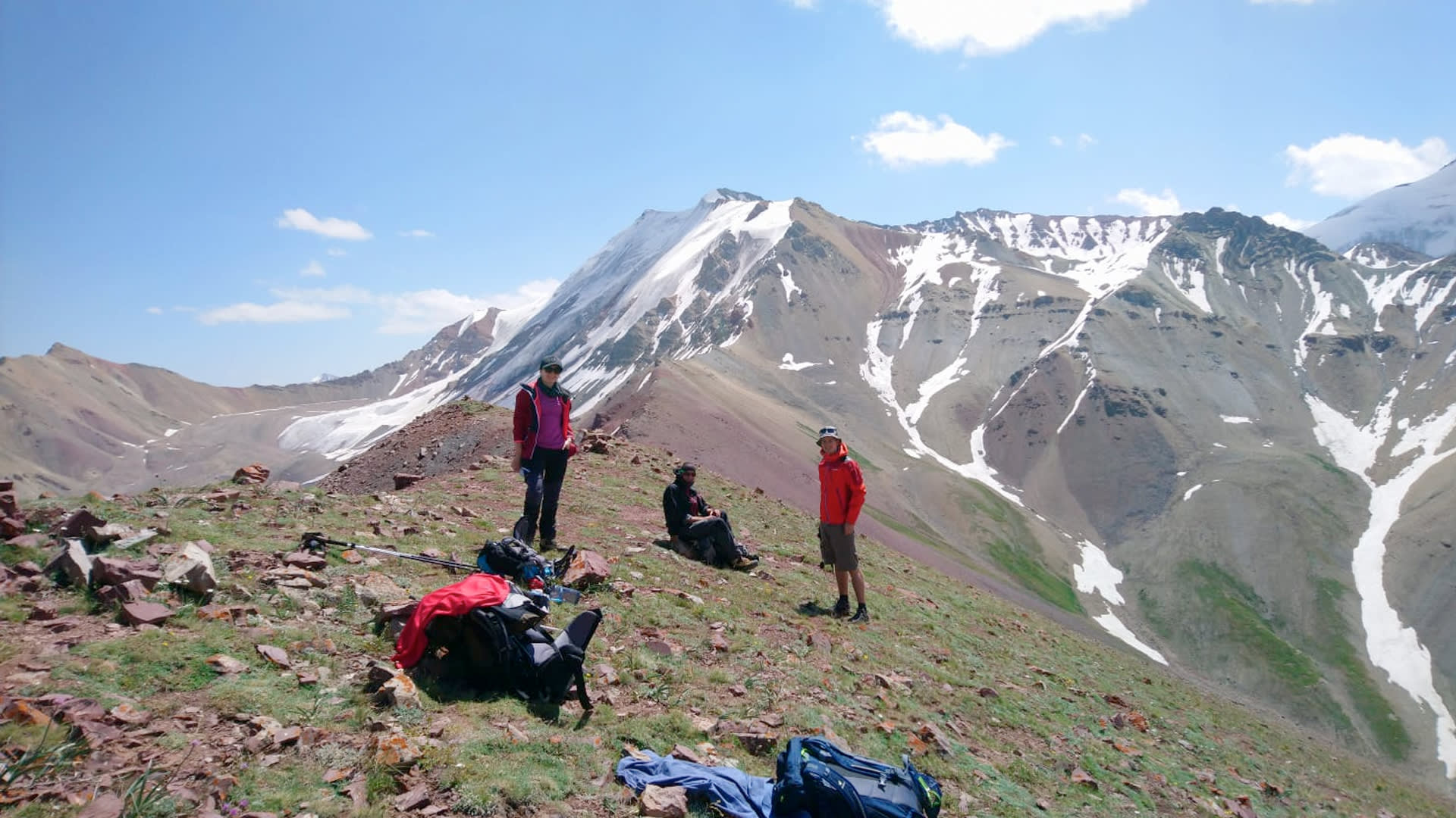 Expedition Pik Lenin: Drei Höhenbergsteiger pausieren auf felsigem Bergrücken, abgelegte Rucksäcke und Wanderstöcke im Vordergrund.