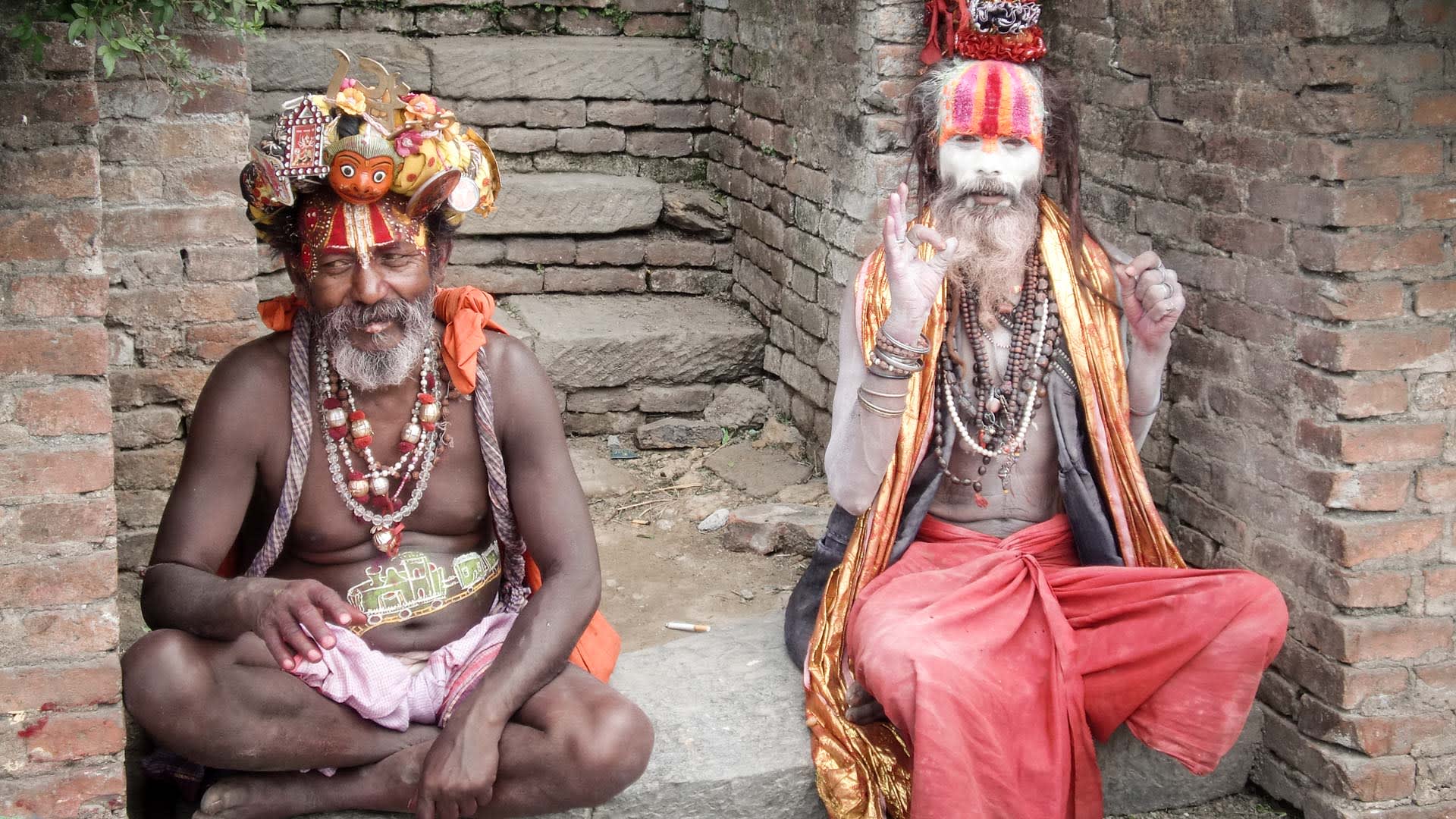 Zwei Sadhus in traditioneller Kleidung und Kopfschmuck sitzen vor einer Backsteinmauer in Pashupatinath.