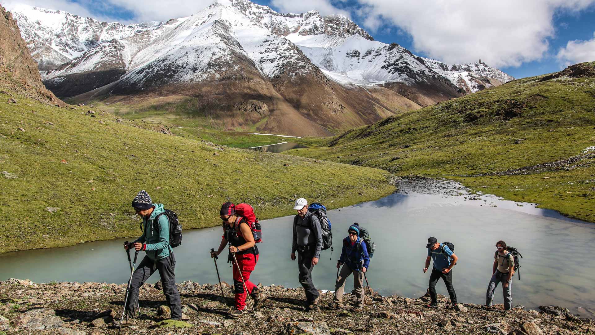 Sechs Höhenbergsteiger mit Trekkingrucksäcken steigen auf steinigem Pfad an zwei Bergseen unterhalb des Pik Lenin auf.