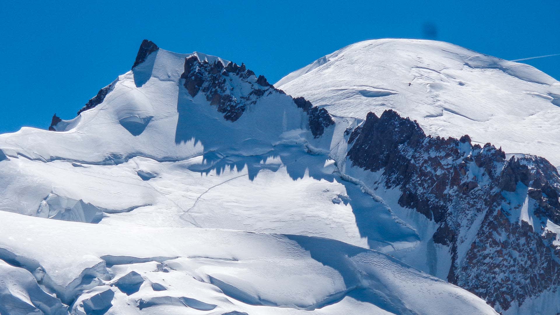 Schmale Aufstiegsspur im Schneefeld am Mont Maudit mit Gletscherspalten und gezacktem Felsgrat im Hintergrund.
