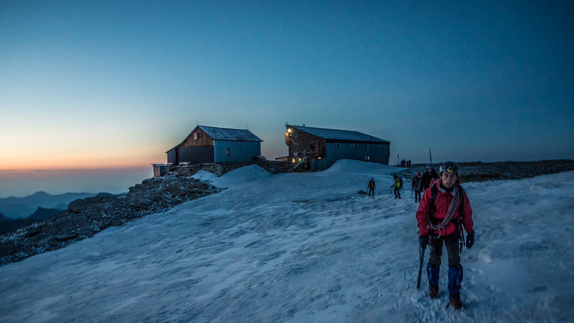 Bergsteiger mit Stirnlampe und Eispickel im Morgengrauen vor der beleuchteten Mantova-Hütte im Wallis.